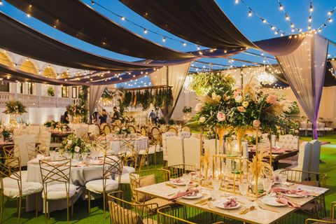 An outdoor evening banquet area with tables, gold chairs, and floral centerpieces under hanging fabric and string lights.