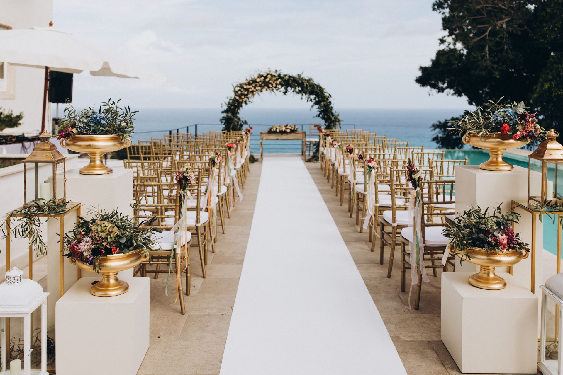 An outdoor wedding aisle lined with chairs, gold pedestals with floral arrangements, and a circular arch facing the ocean. An outdoor wedding aisle lined with chairs, gold pedestals with floral arrangements, and a circular arch facing the ocean.
