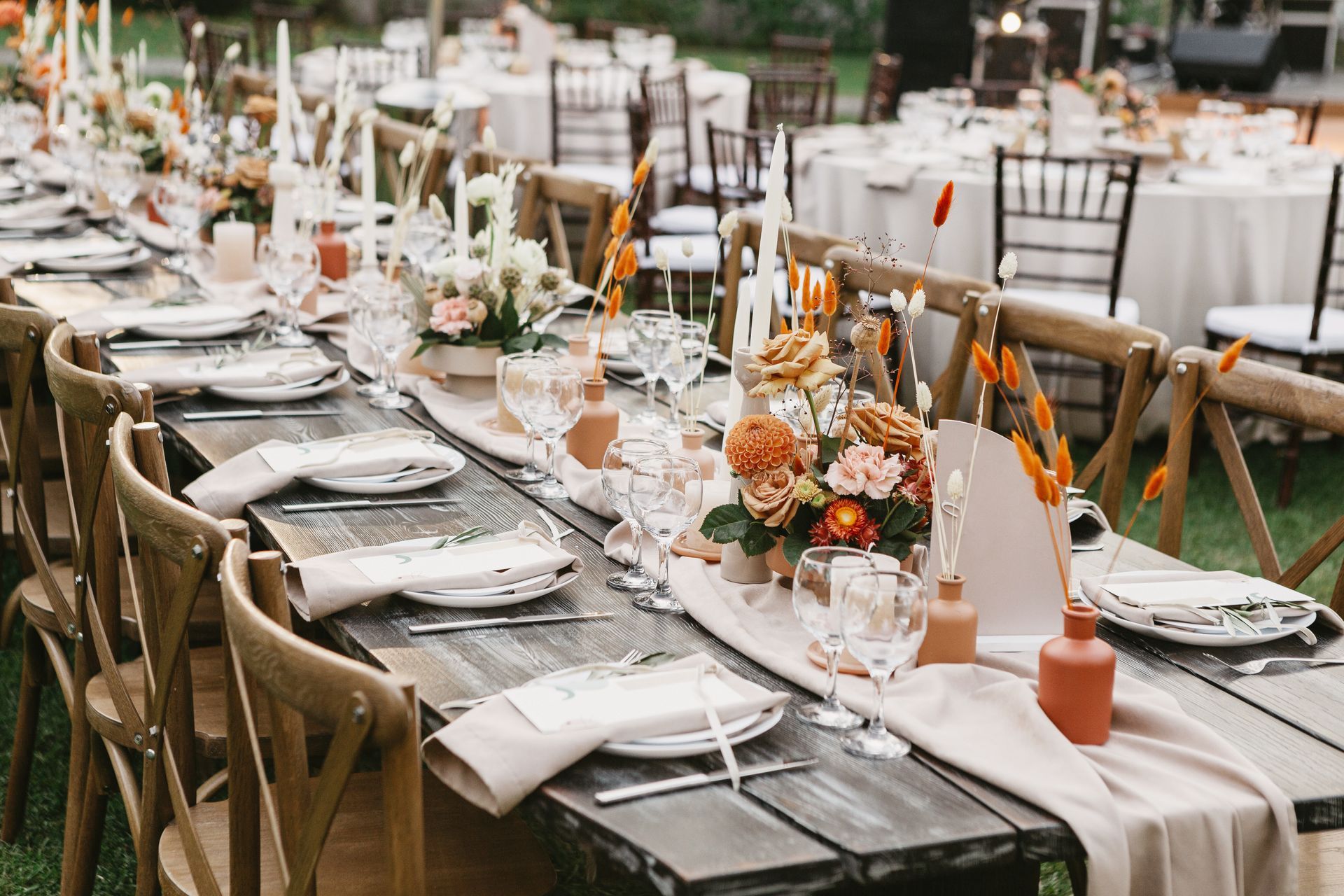 A long outdoor wedding banquet table with wooden chairs, decorated with floral arrangements, candles, and linens.