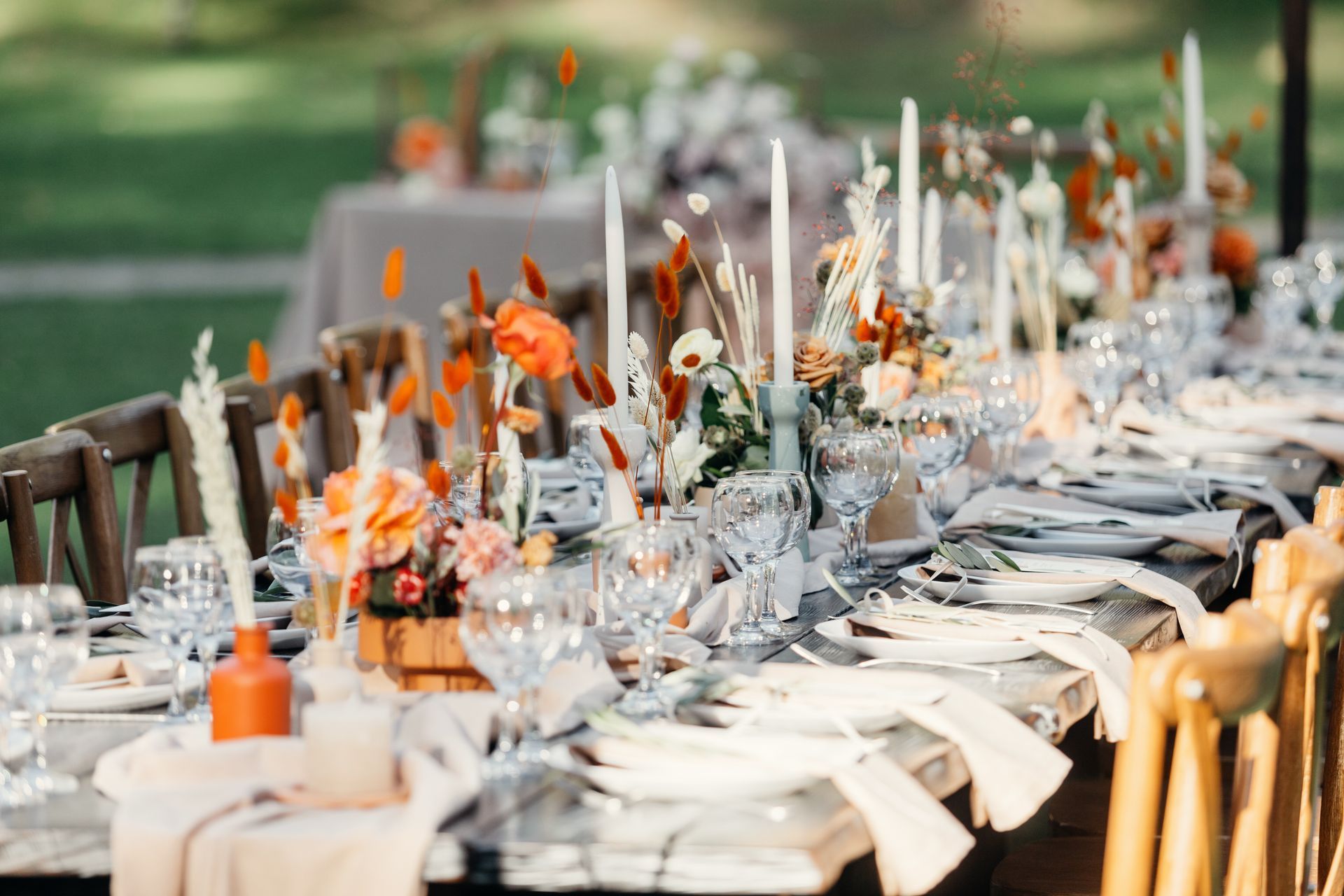 A long table set for an outdoor wedding with orange florals, white tapered candles, and light table linens. A long table set for an outdoor wedding with orange florals, white tapered candles, and light table linens.