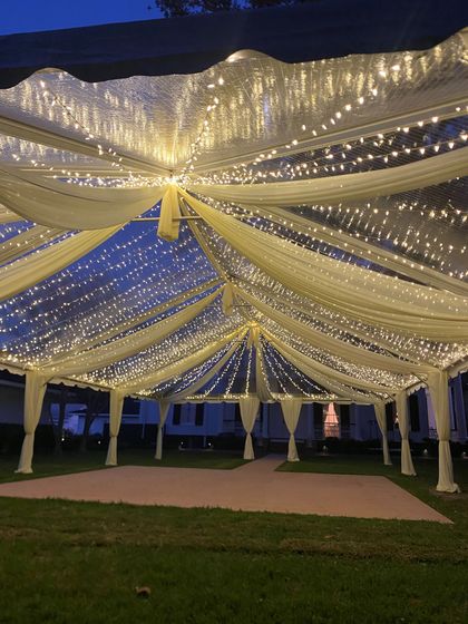 Draped white fabric canopy lit with warm string lights over a grassy outdoor event space at night