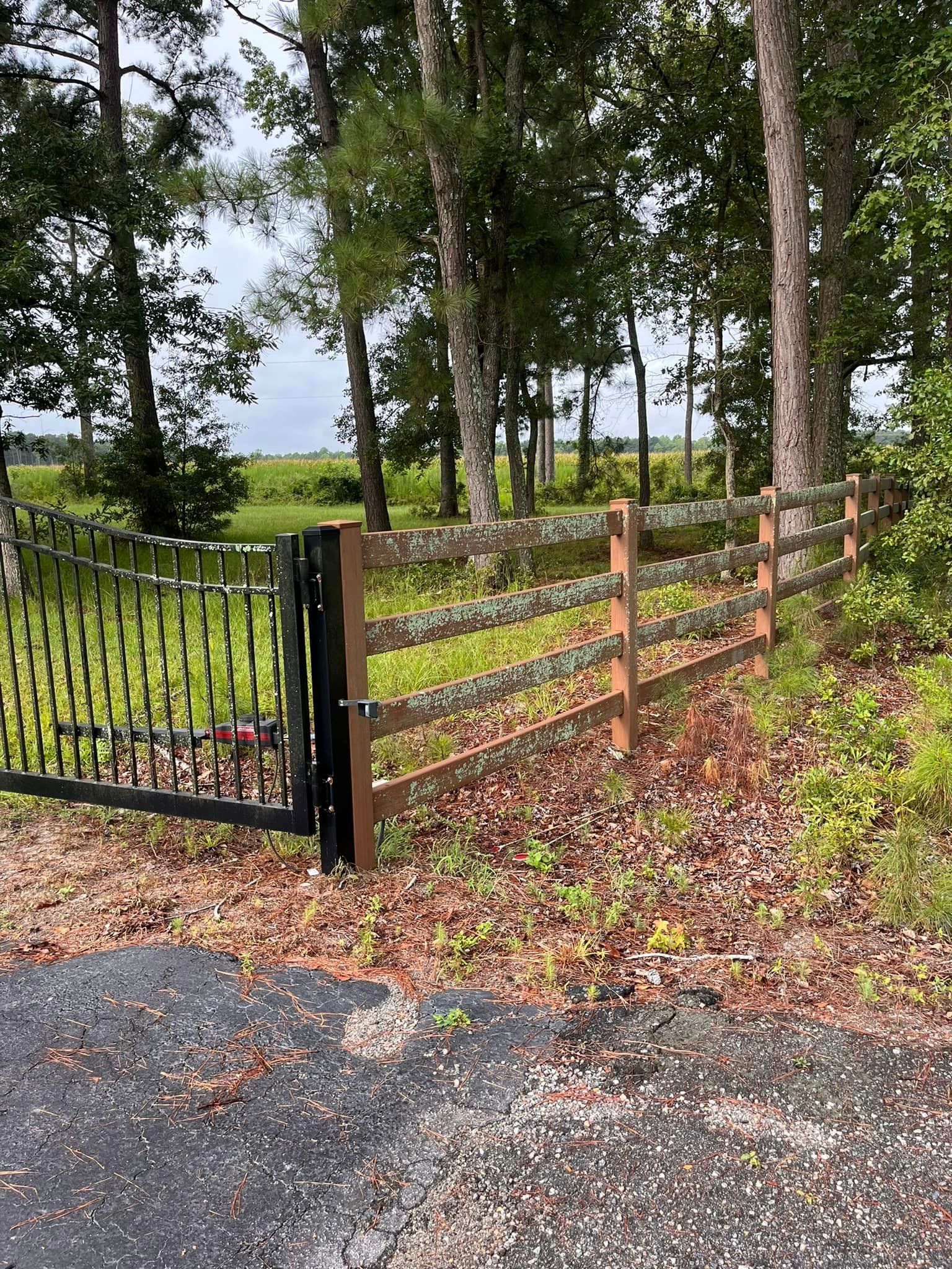 A wooden fence with a metal gate is surrounded by trees on the side of a road.