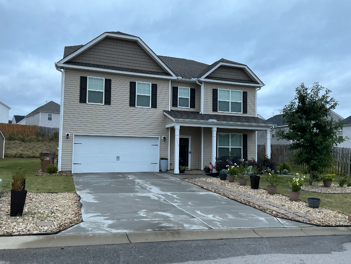 Suburban two-story beige house with attached garage, stone accents, and a concrete driveway under cloudy sky