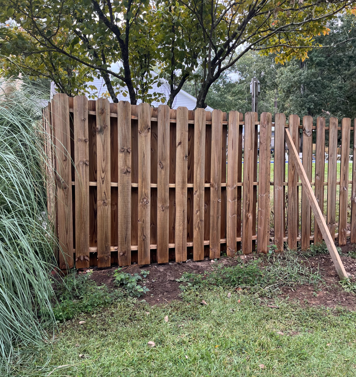 Wooden backyard fence with a leaning post, grass in front, and trees behind