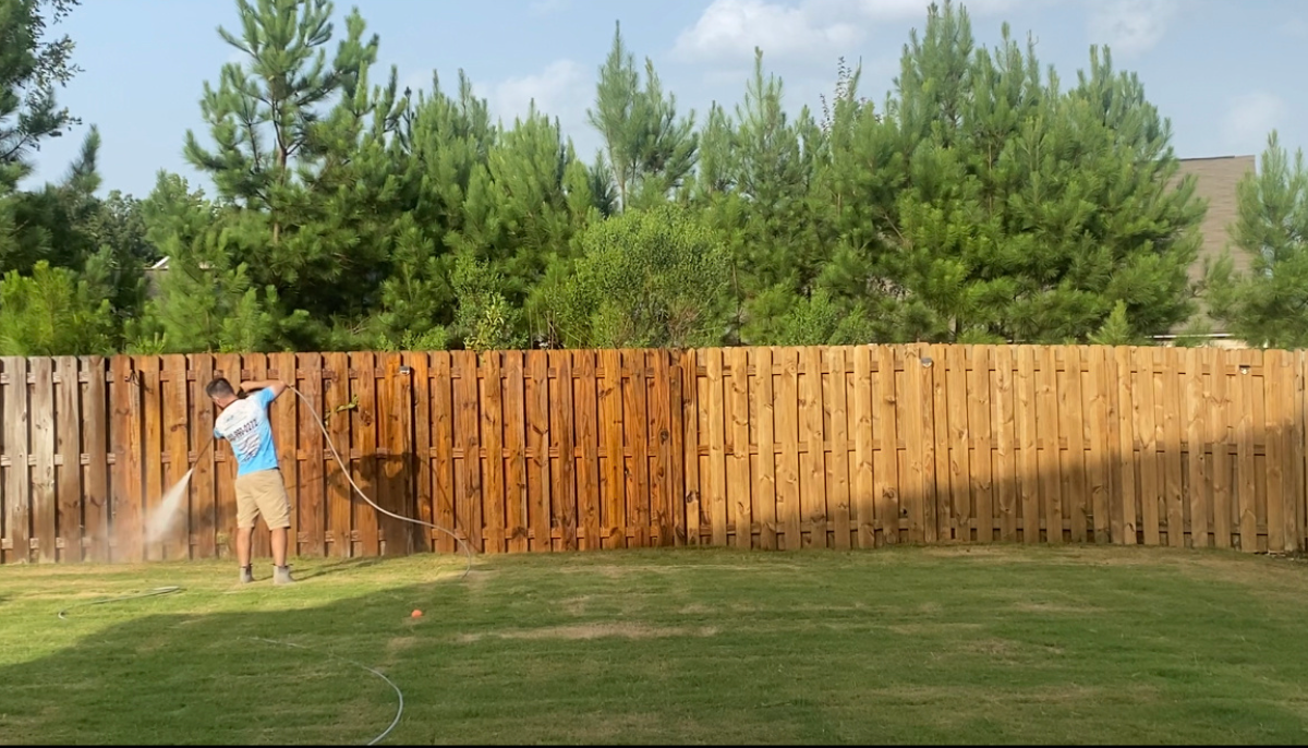 Person painting a long wooden fence brown in a grassy yard with trees behind it.