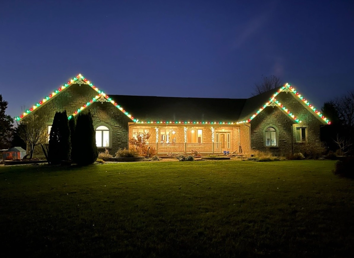 Single-story house at night, outlined with colorful Christmas lights and lit windows, on a dark lawn.