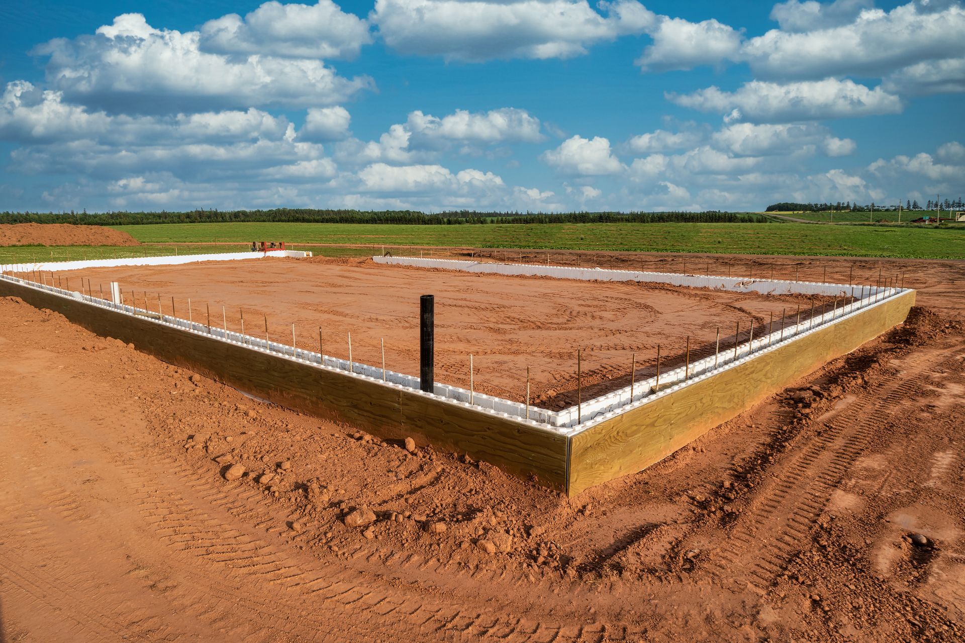 Foundation of a building under construction on red dirt with a blue sky and clouds in the background.