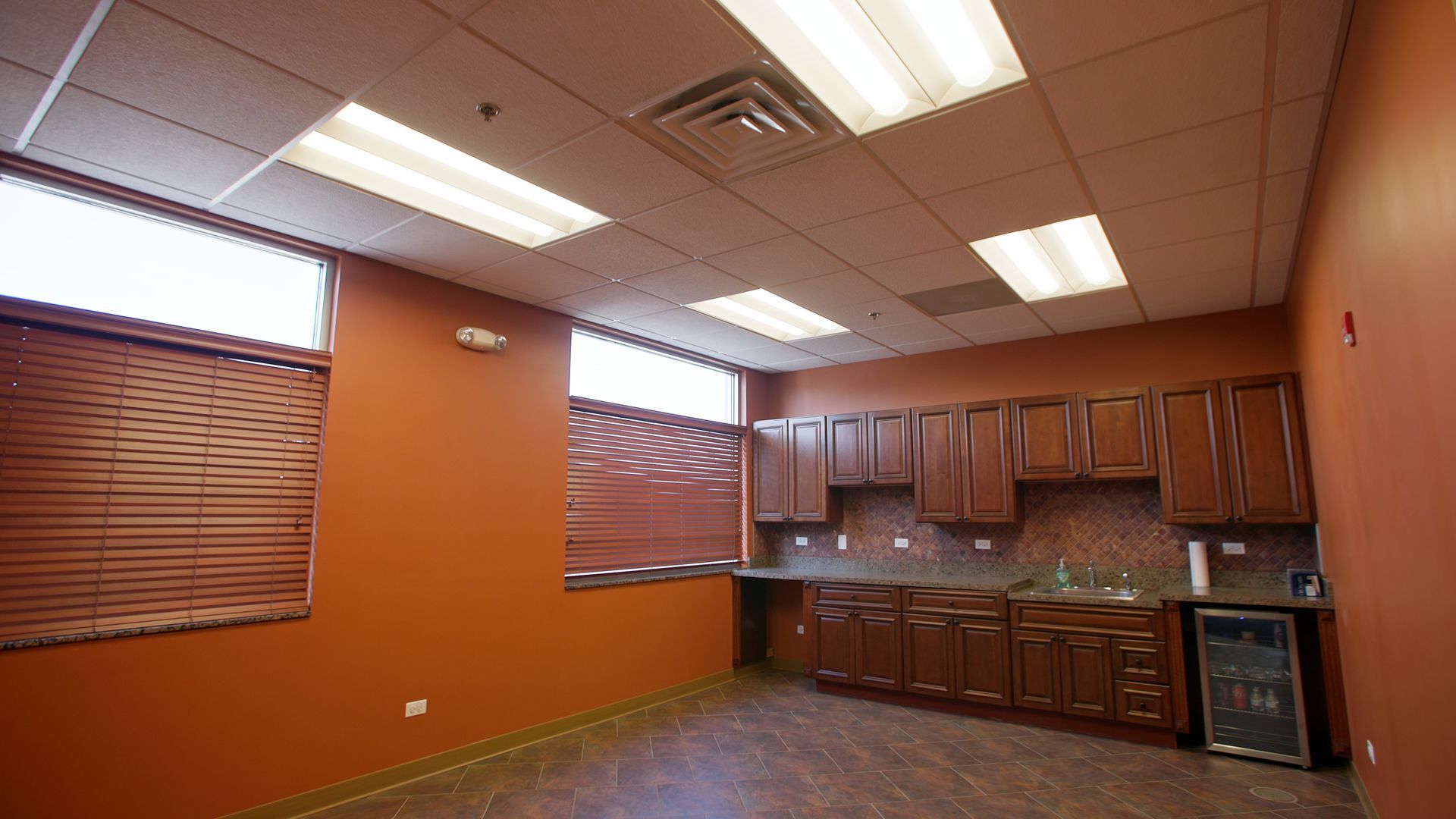 Empty office break room with orange walls, wood cabinets, sink, mini fridge, and fluorescent ceiling lights