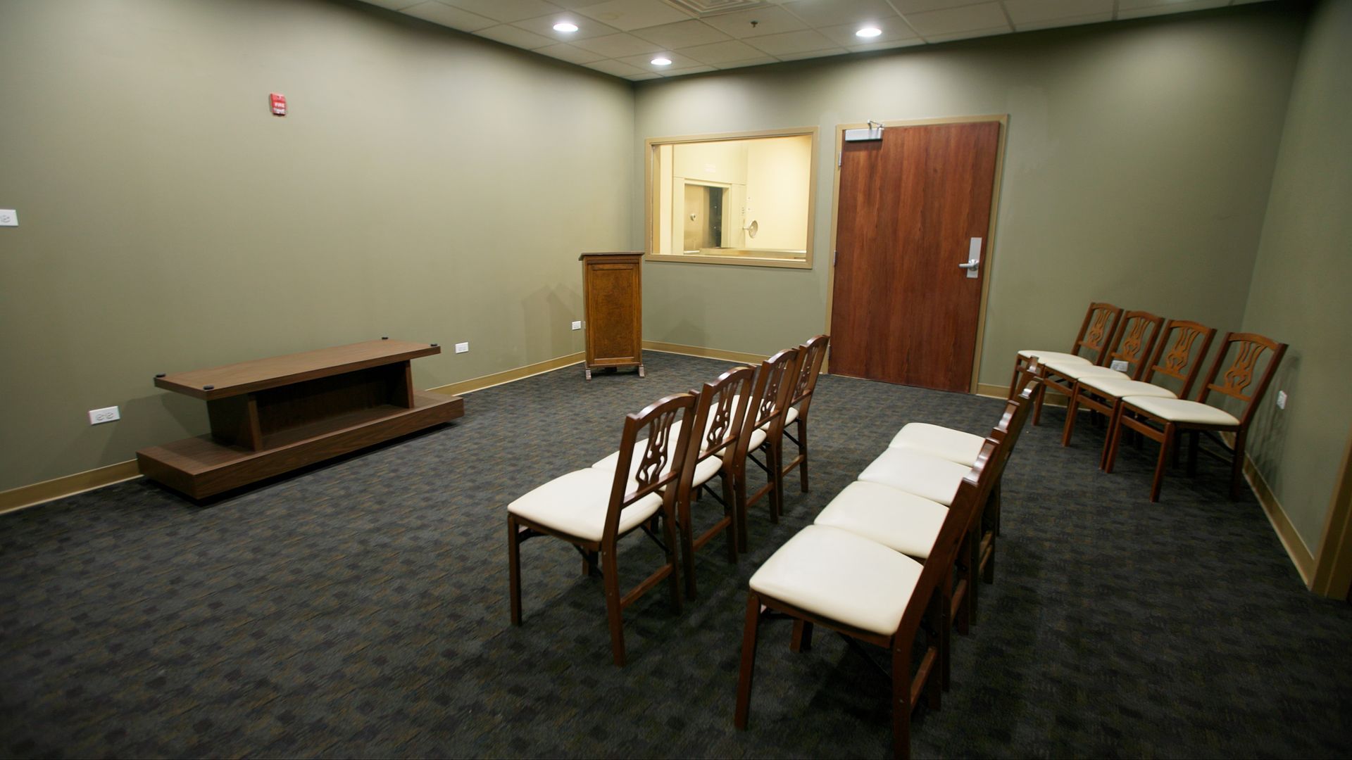 Empty waiting room with rows of chairs, beige walls, a carpeted floor, and a wooden door at the back.