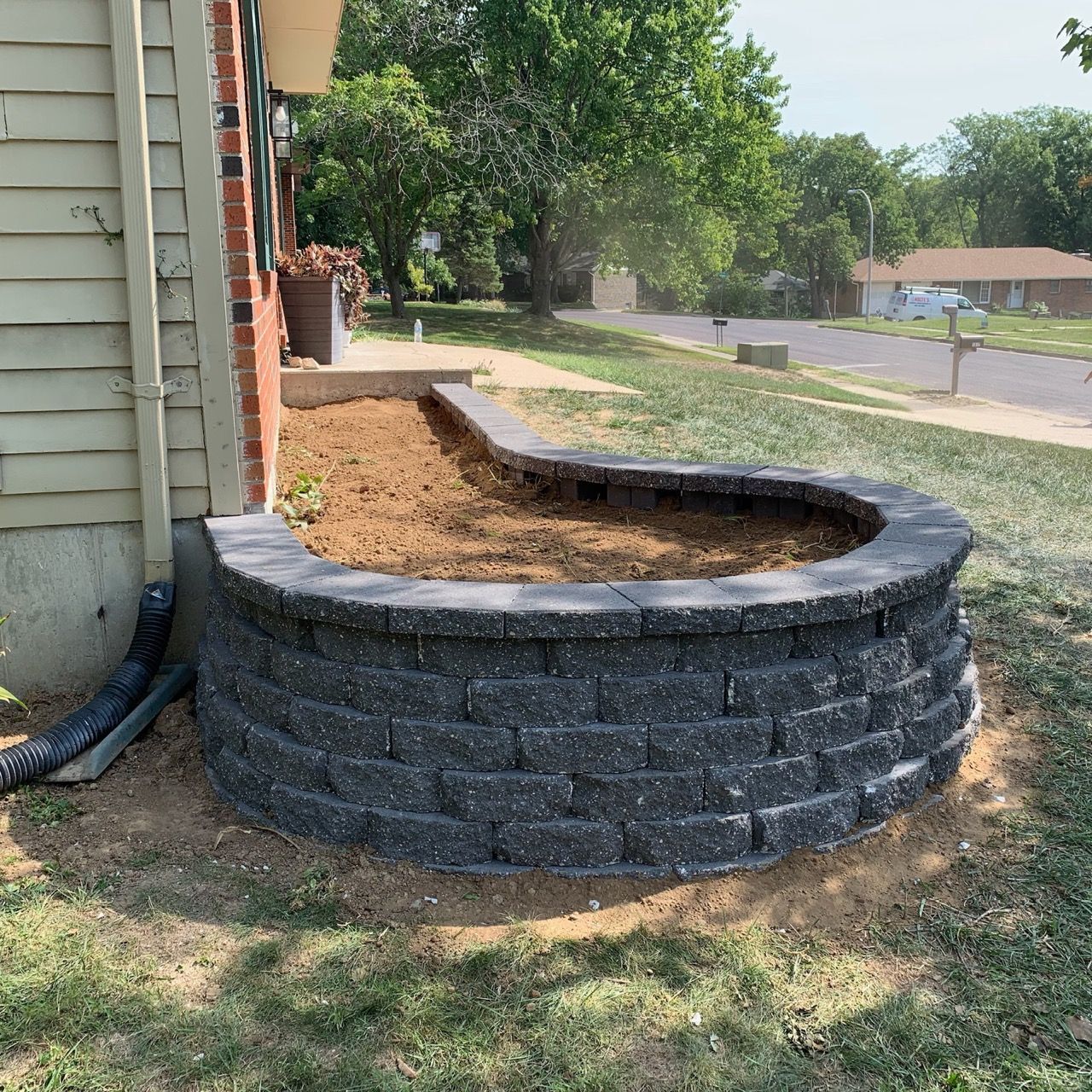 Raised brick garden bed beside a house, with bare soil and curved dark retaining wall under construction