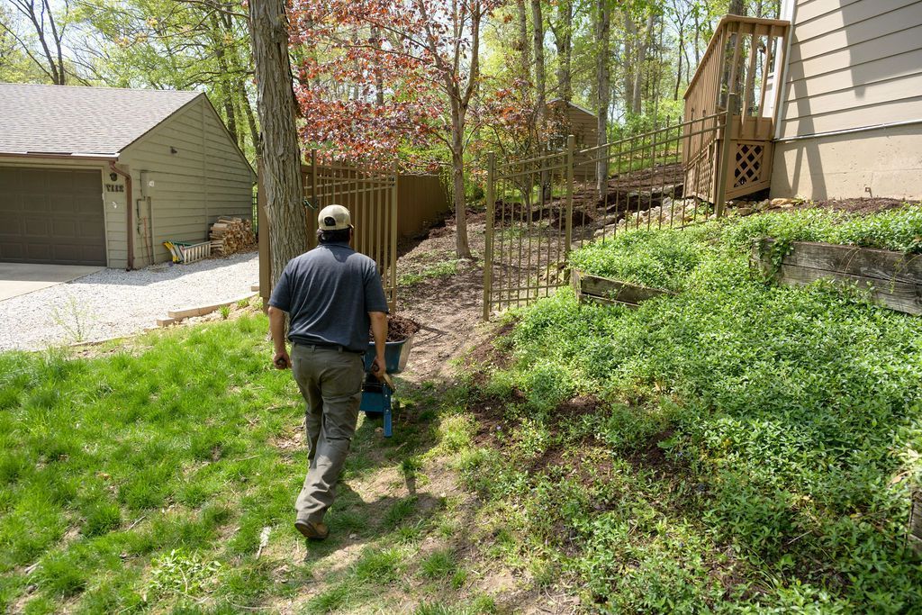 Person walking up a grassy slope beside houses and trees, carrying a blue bucket.
