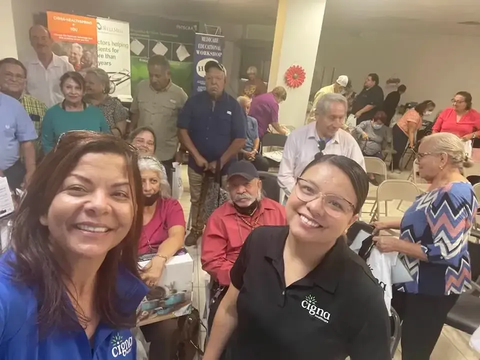 Smiling group selfie at an indoor gathering, with people seated and standing in the background.