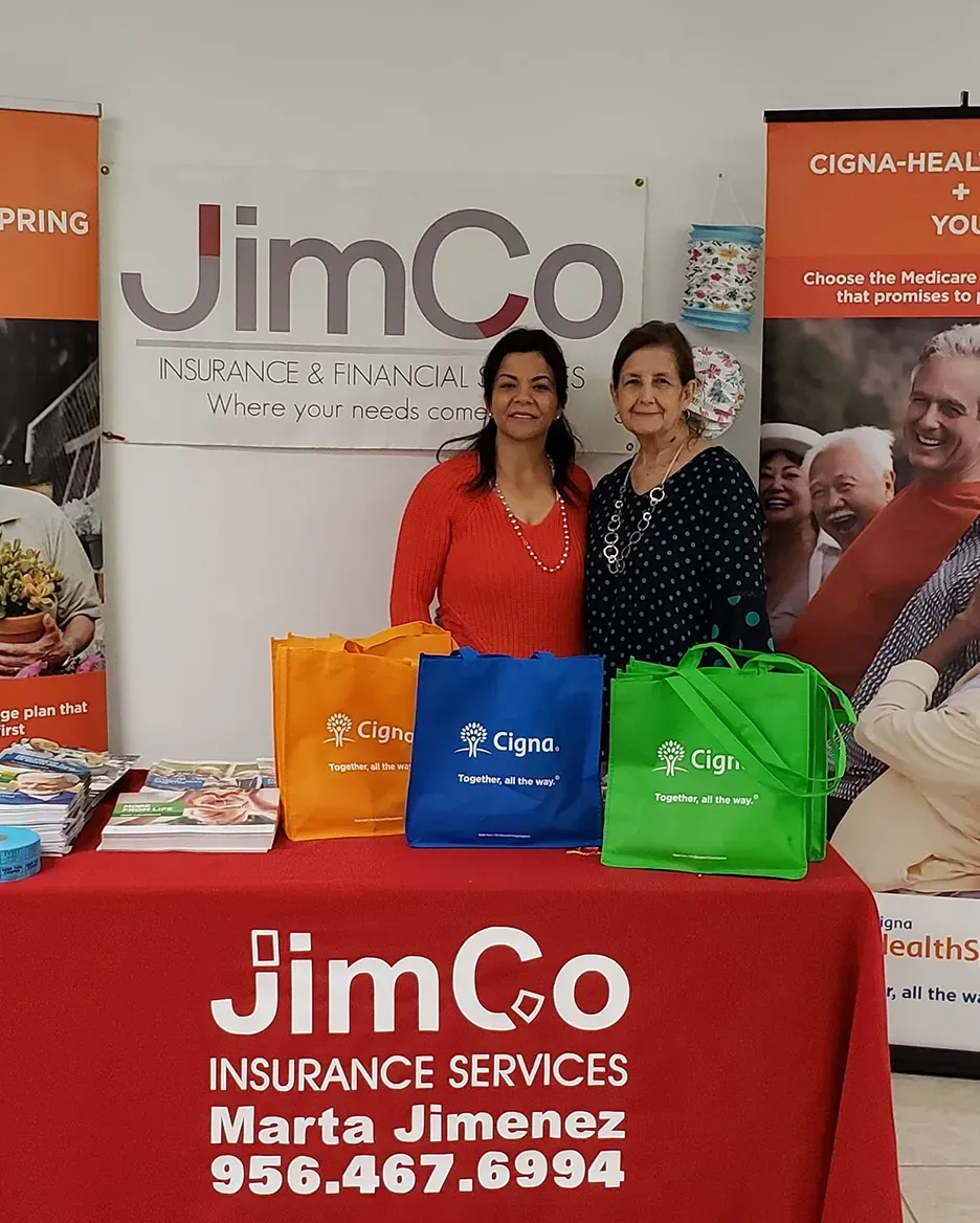 Two women behind a JimCo insurance services booth with colorful bags and banners