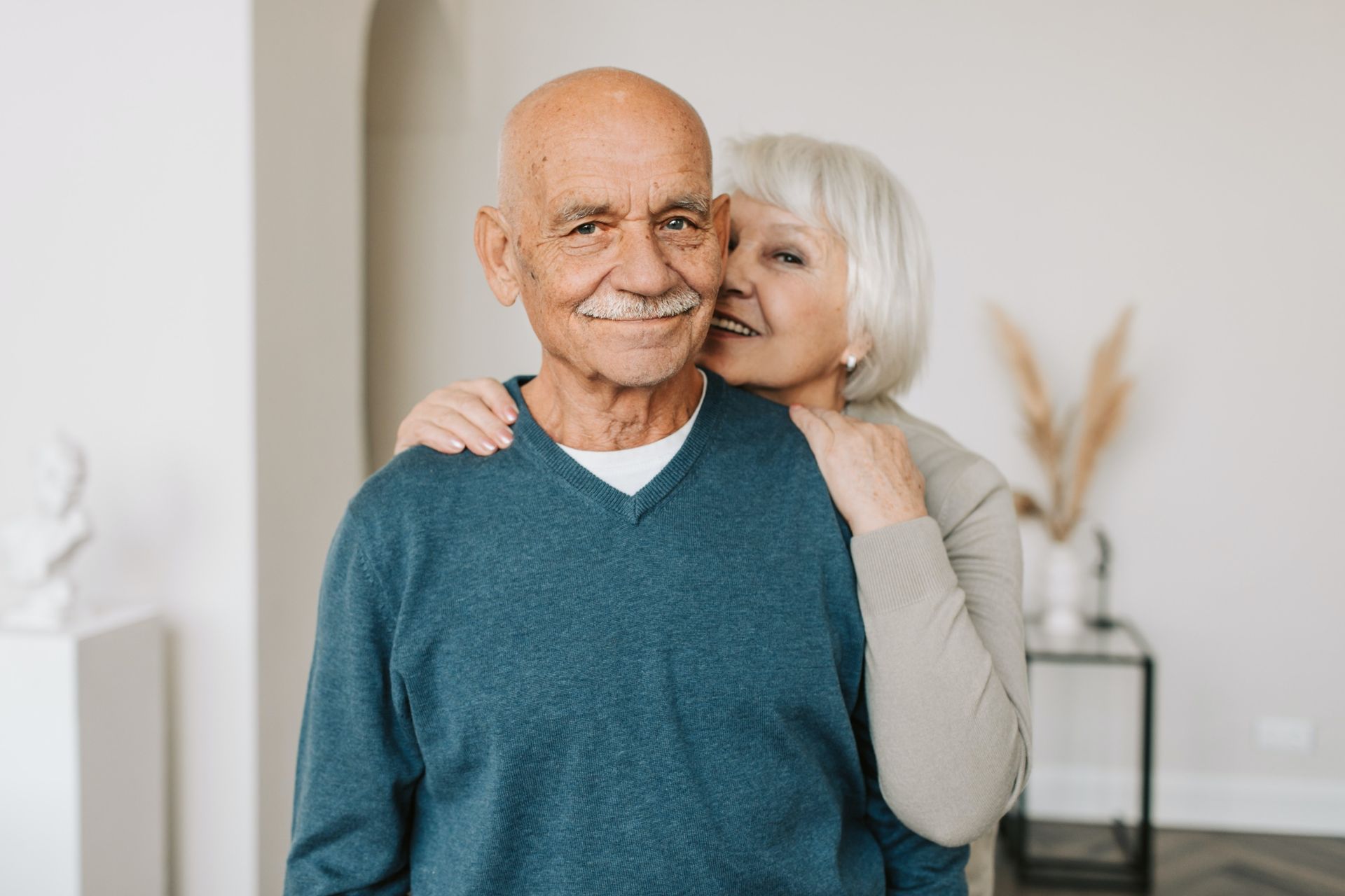 Older couple in a bright home, smiling and embracing near a window.