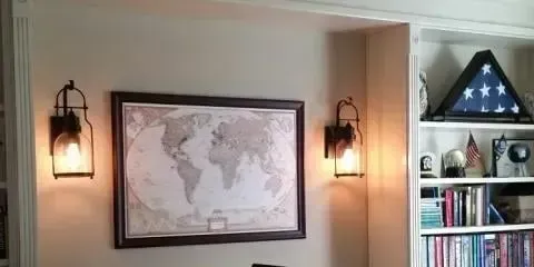 A wall featuring a framed world map flanked by two wall-mounted lanterns, next to a bookcase filled with items.