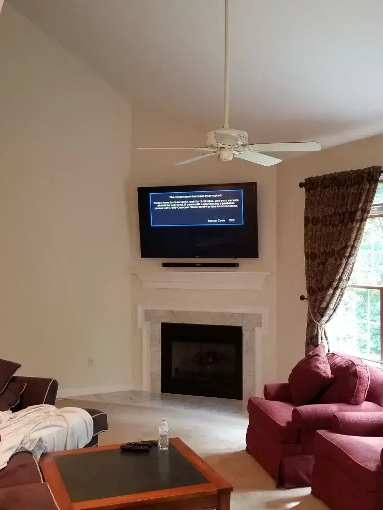 A TV mounted above a fireplace in a living room with a ceiling fan, a dark coffee table, and maroon armchairs.