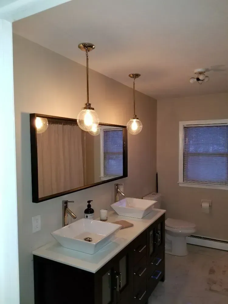 A bathroom vanity with a long black-framed mirror, two pendant lights, and two square sinks over a dark wood cabinet.