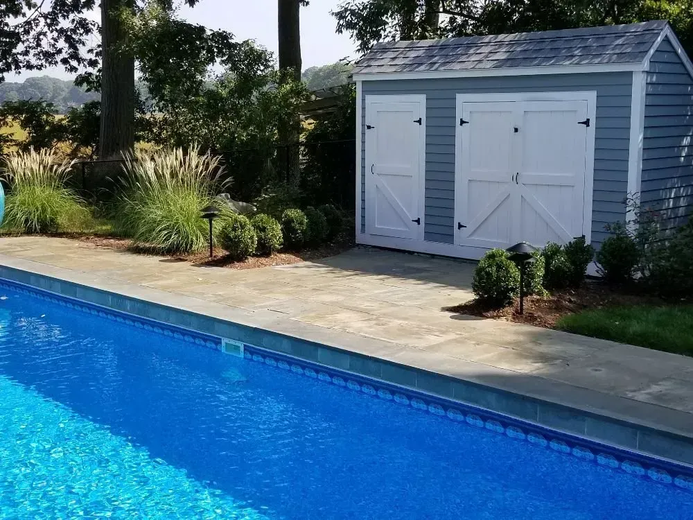 A blue swimming pool sits next to a light blue storage shed with white doors in a landscaped backyard.