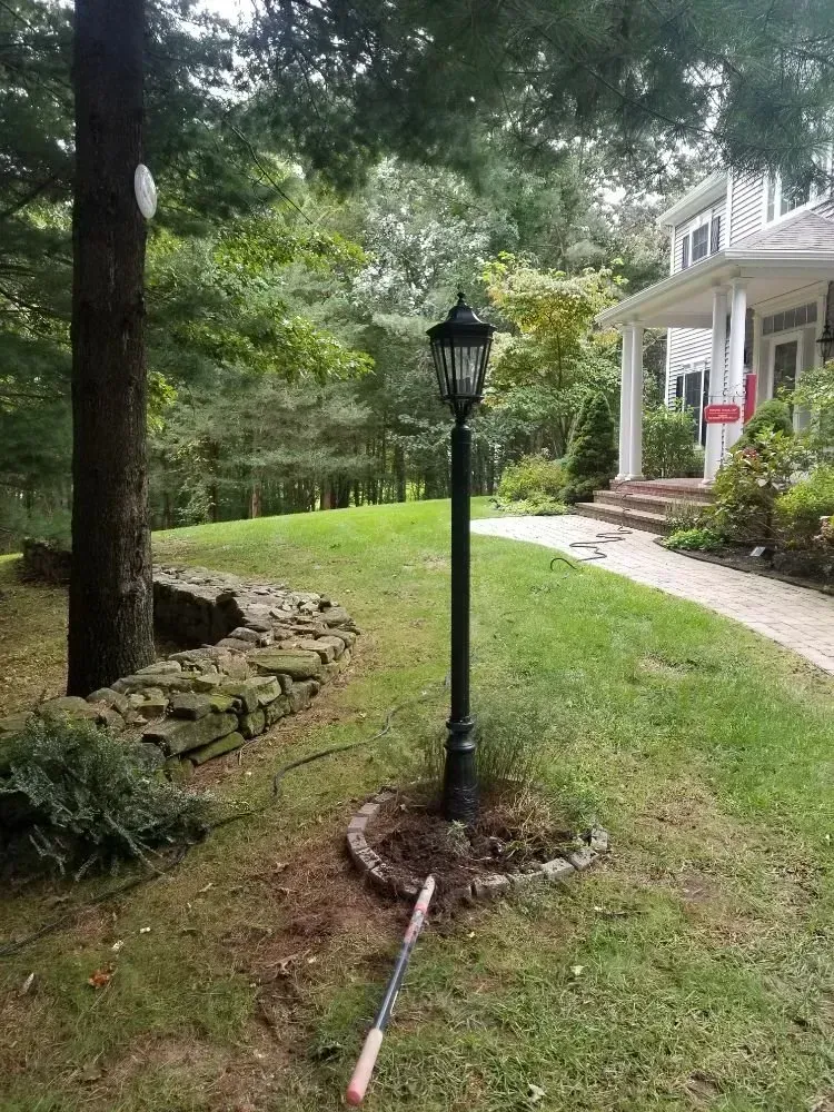 A black post lamp stands in a small, circular garden bed on a lawn, with a house and stone wall in the background.