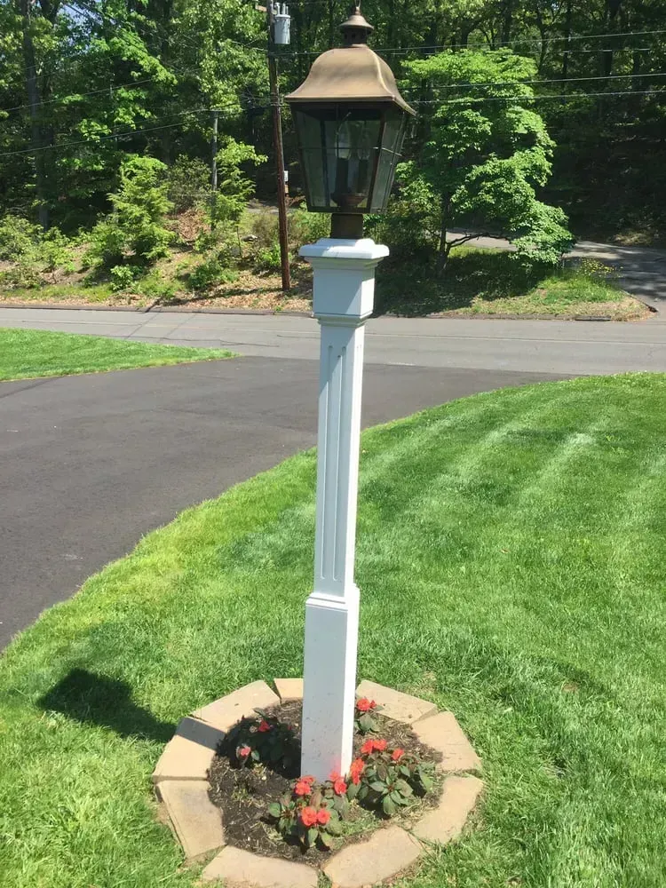 A white lamp post stands in a circular flower bed with red flowers, set in a grassy yard next to a paved driveway.