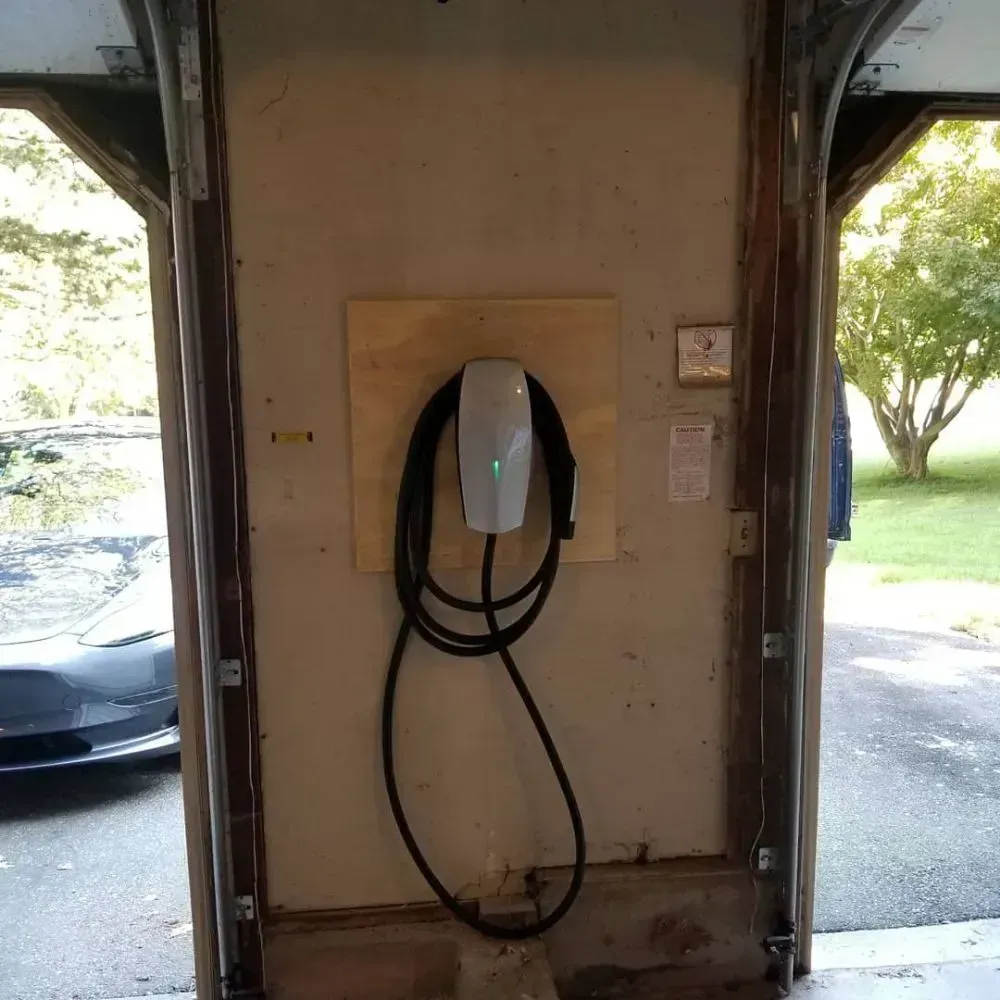 A Tesla Wall Connector mounted on a wooden board on a garage wall, with its charging cable coiled around it.
