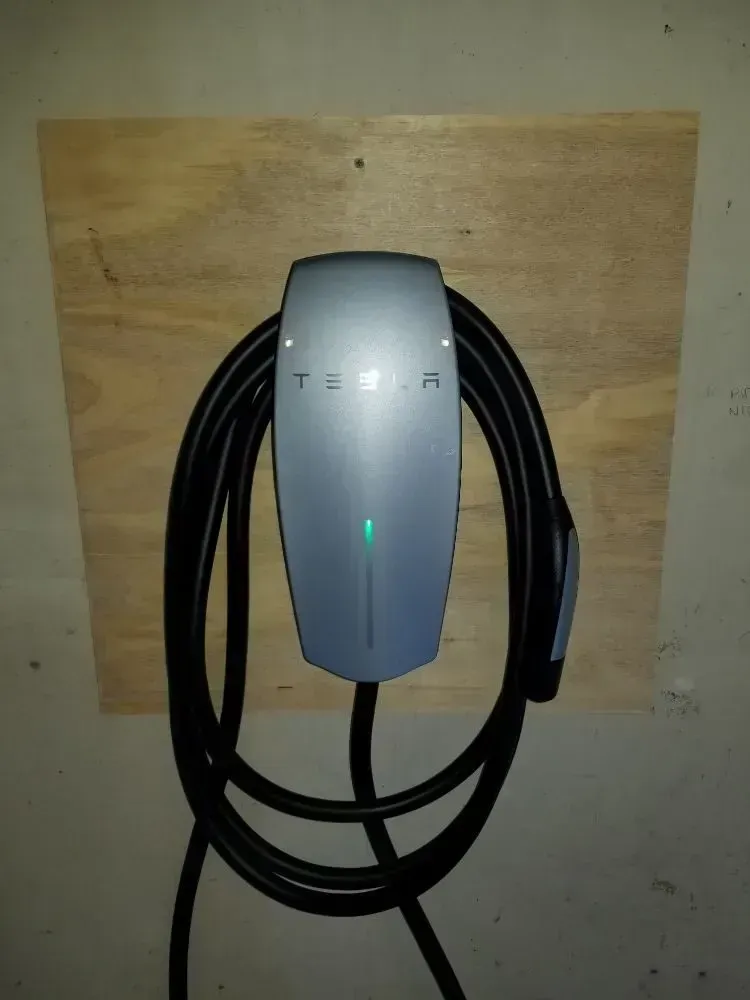 A silver Tesla Wall Connector mounted on a wooden board against a wall, with the charging cable coiled and light on.