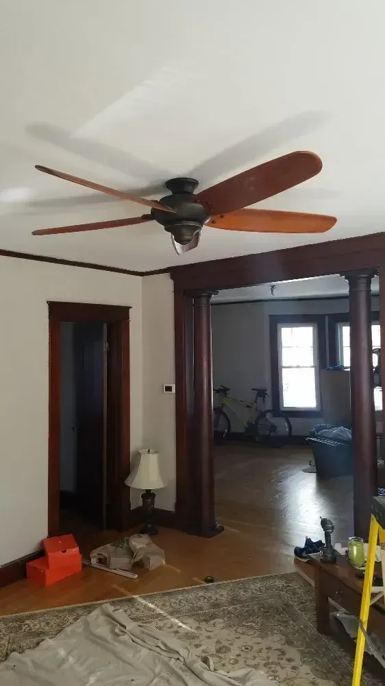 Ceiling fan in a living room with dark wood trim, leading to a second room with bicycles and hardwood floors.