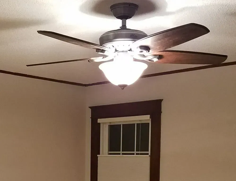 A dark brown ceiling fan with a bright, glowing glass light fixture installed in a room with dark wood window trim.