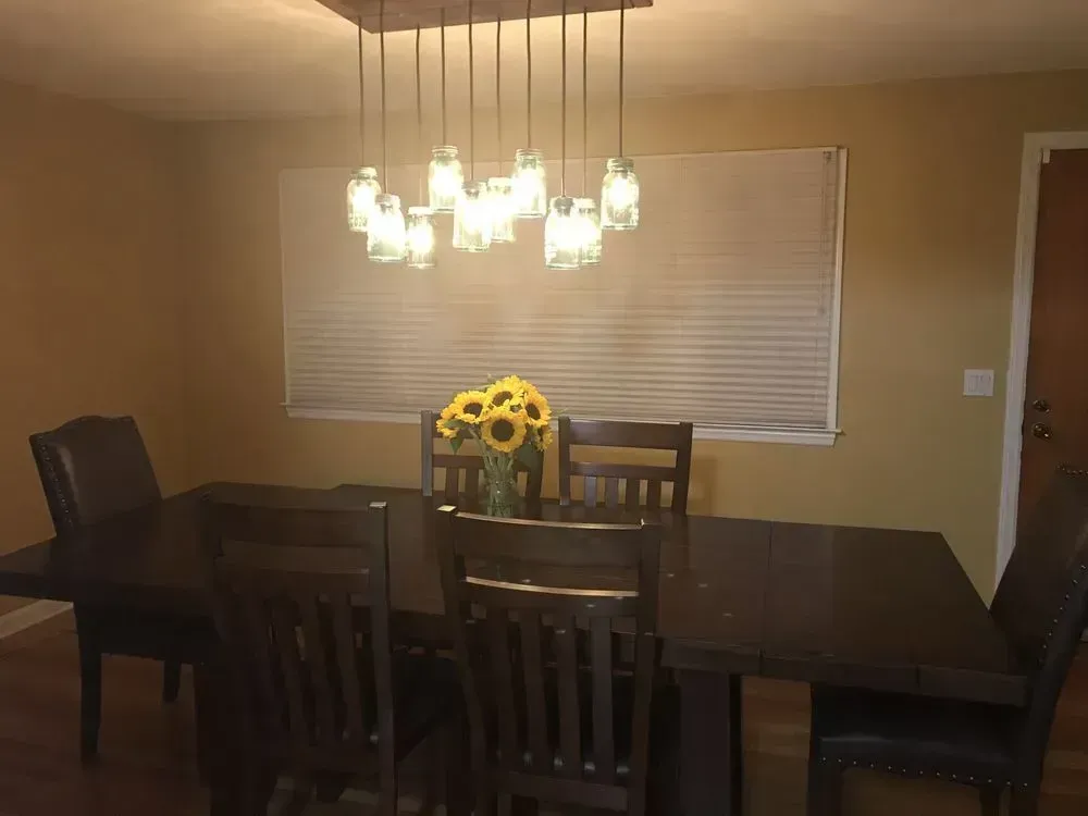 A dark wood dining table and chairs set under a multi-pendant mason jar light fixture, with sunflowers in the center.
