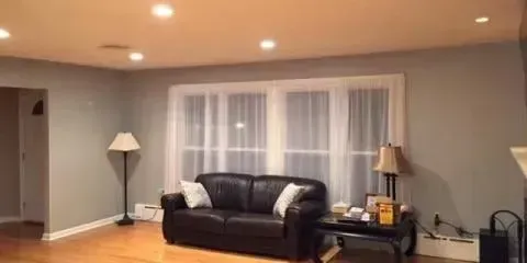 A living room with gray walls, a black leather sofa against a window with white curtains, and a floor lamp on the left.
