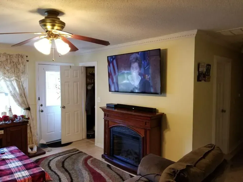 A living room featuring a mounted television above a fireplace, a ceiling fan, and an open door to the outside.