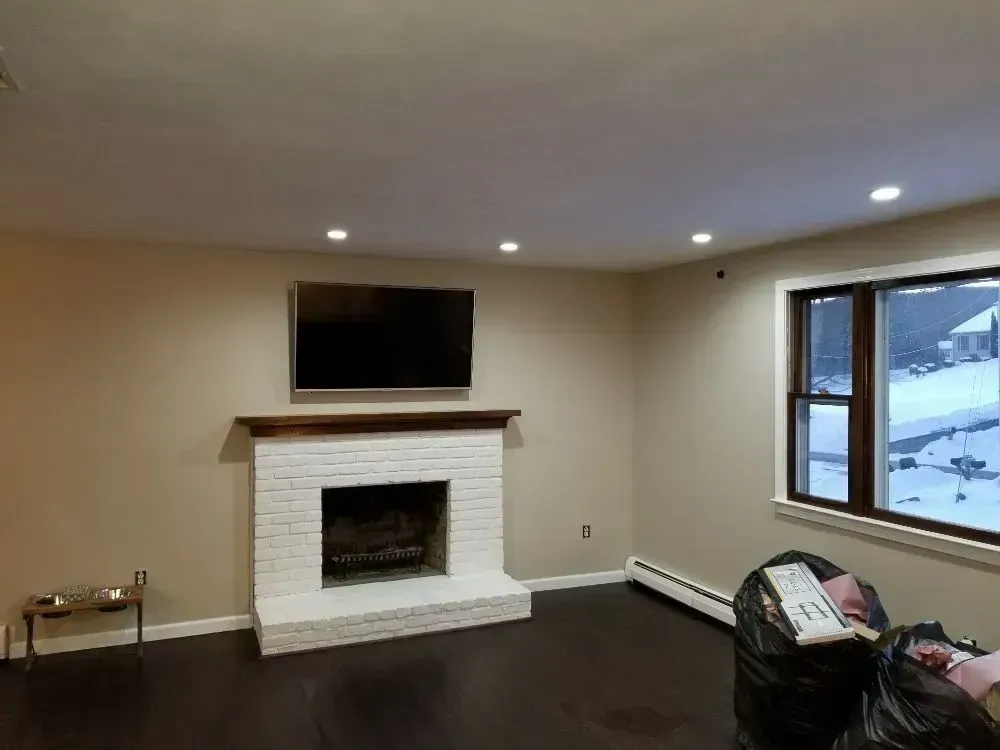 A room with a white brick fireplace, mounted television, dark hardwood floors, and a window overlooking a snowy yard.