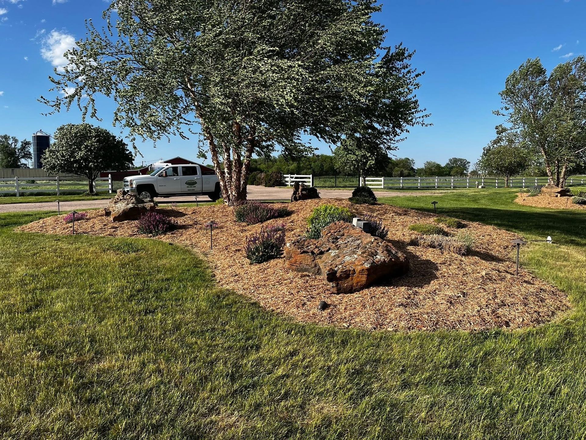 Landscaped area with rocks, mulch, plants, and a tree on a green lawn with a truck in the background.