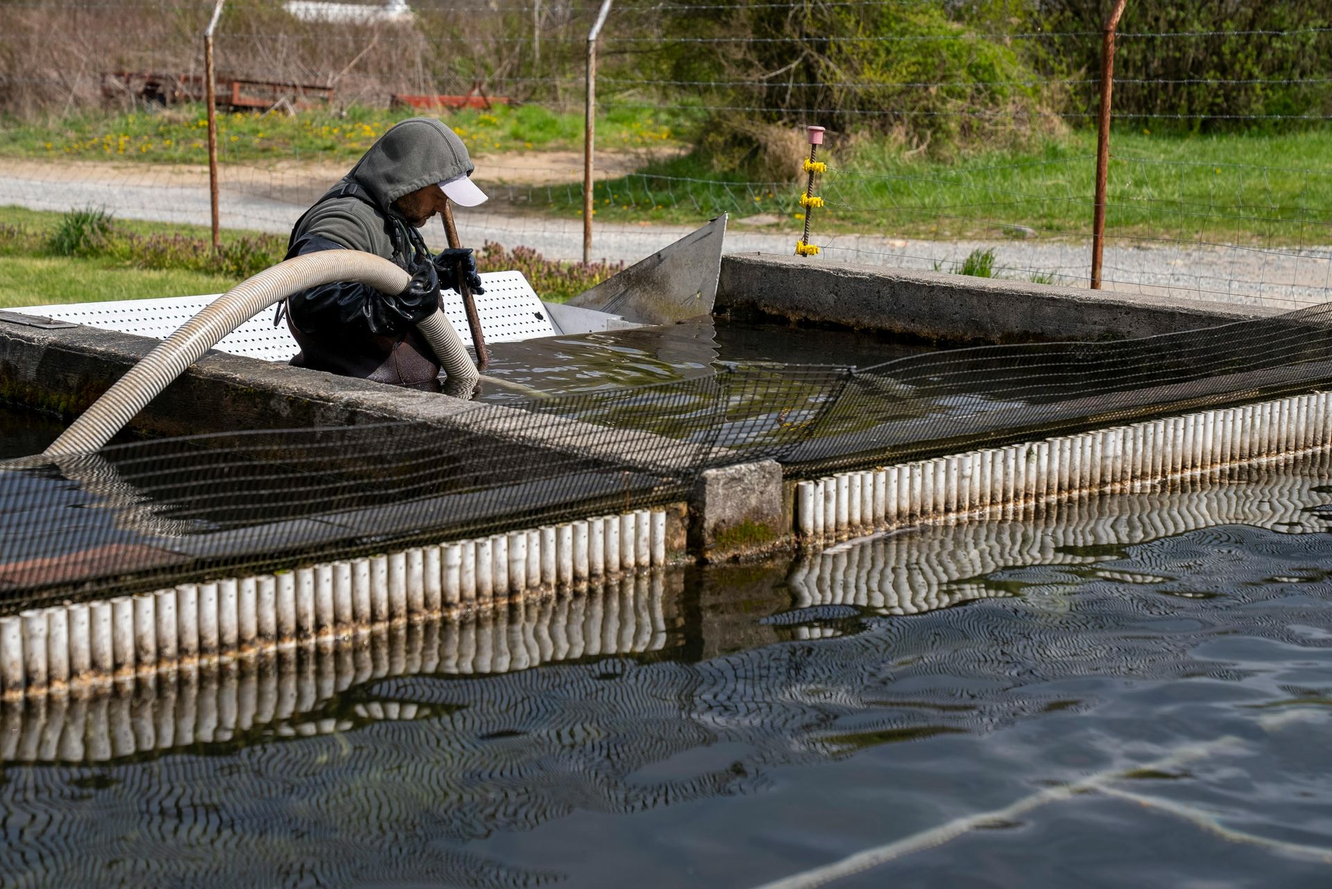 Person in hooded jacket cleans a water-filled tank with a hose and a net, outdoors.