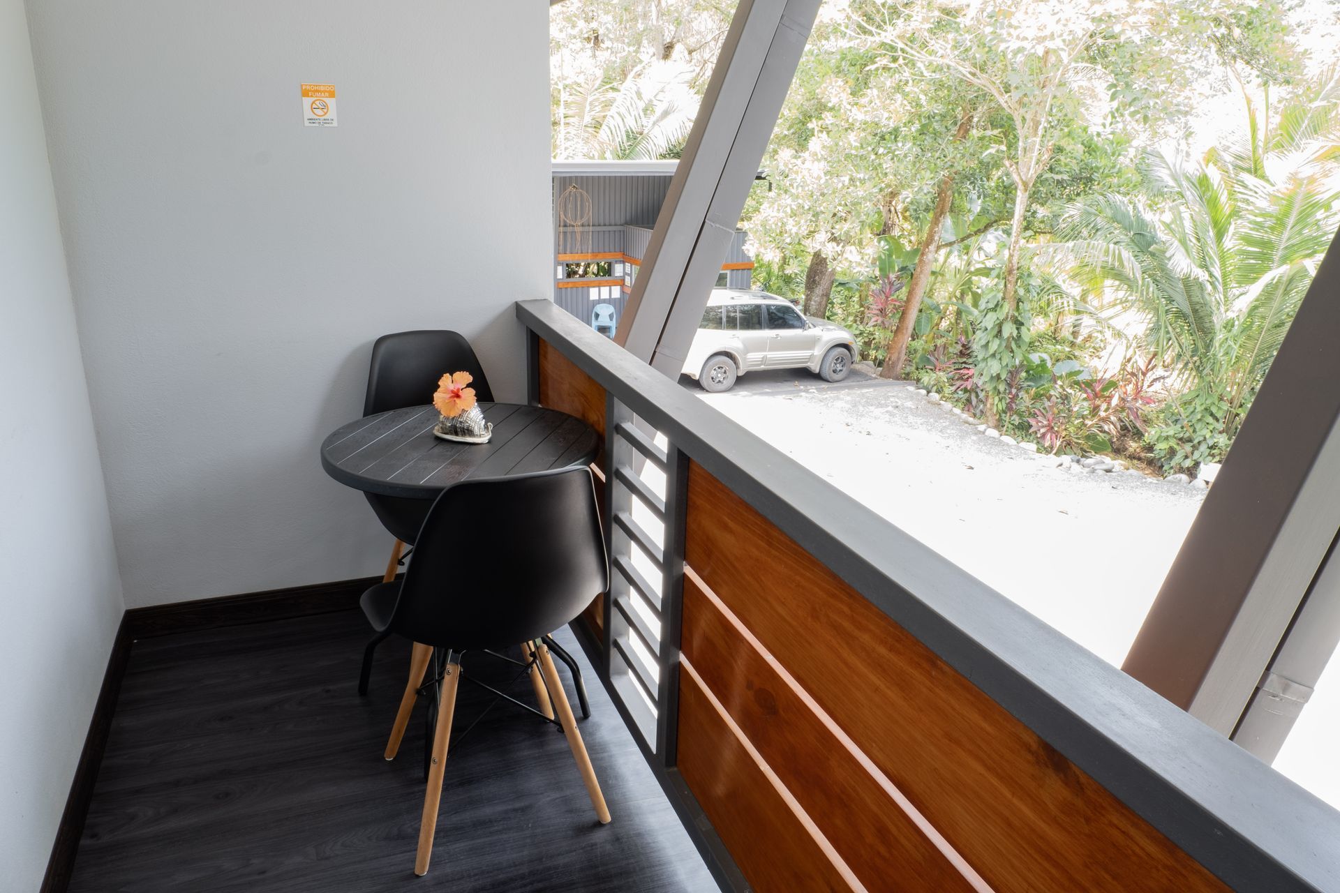 A balcony with a small round black table and two matching chairs, overlooking a driveway with a parked white SUV.