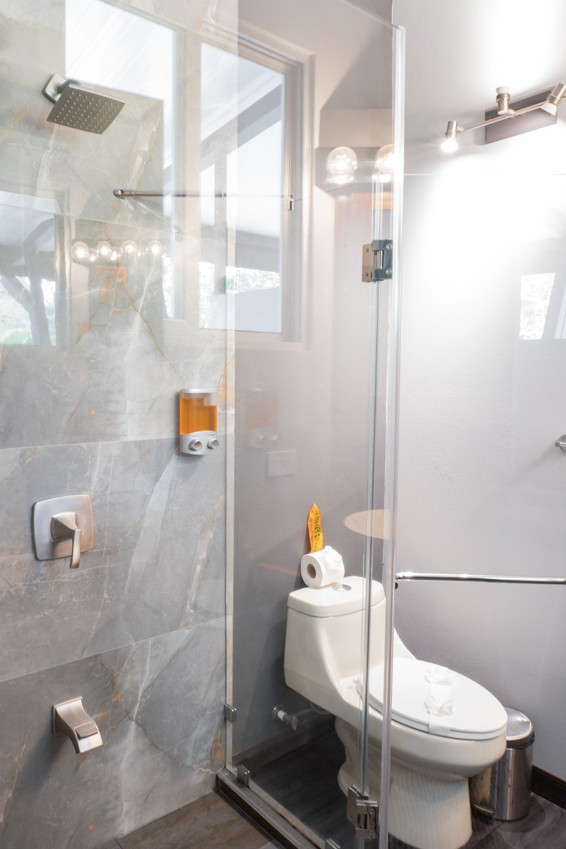 A modern, neutral-toned bathroom featuring a walk-in shower with stone-look tiles next to a white toilet with a grab bar.