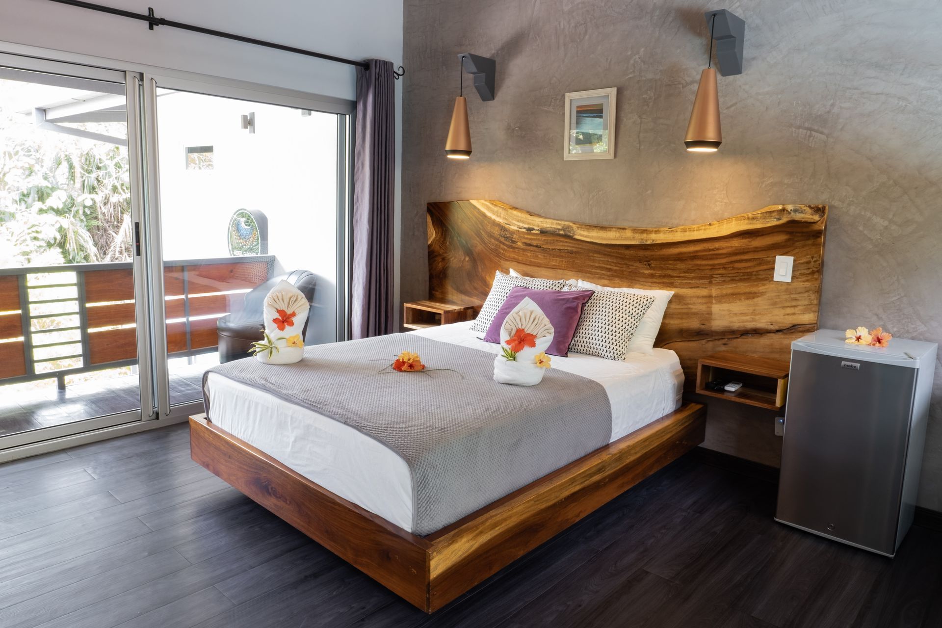 A modern bedroom with a live-edge wooden headboard, a gray bed, two pendant lights, and a sliding glass door to a balcony.