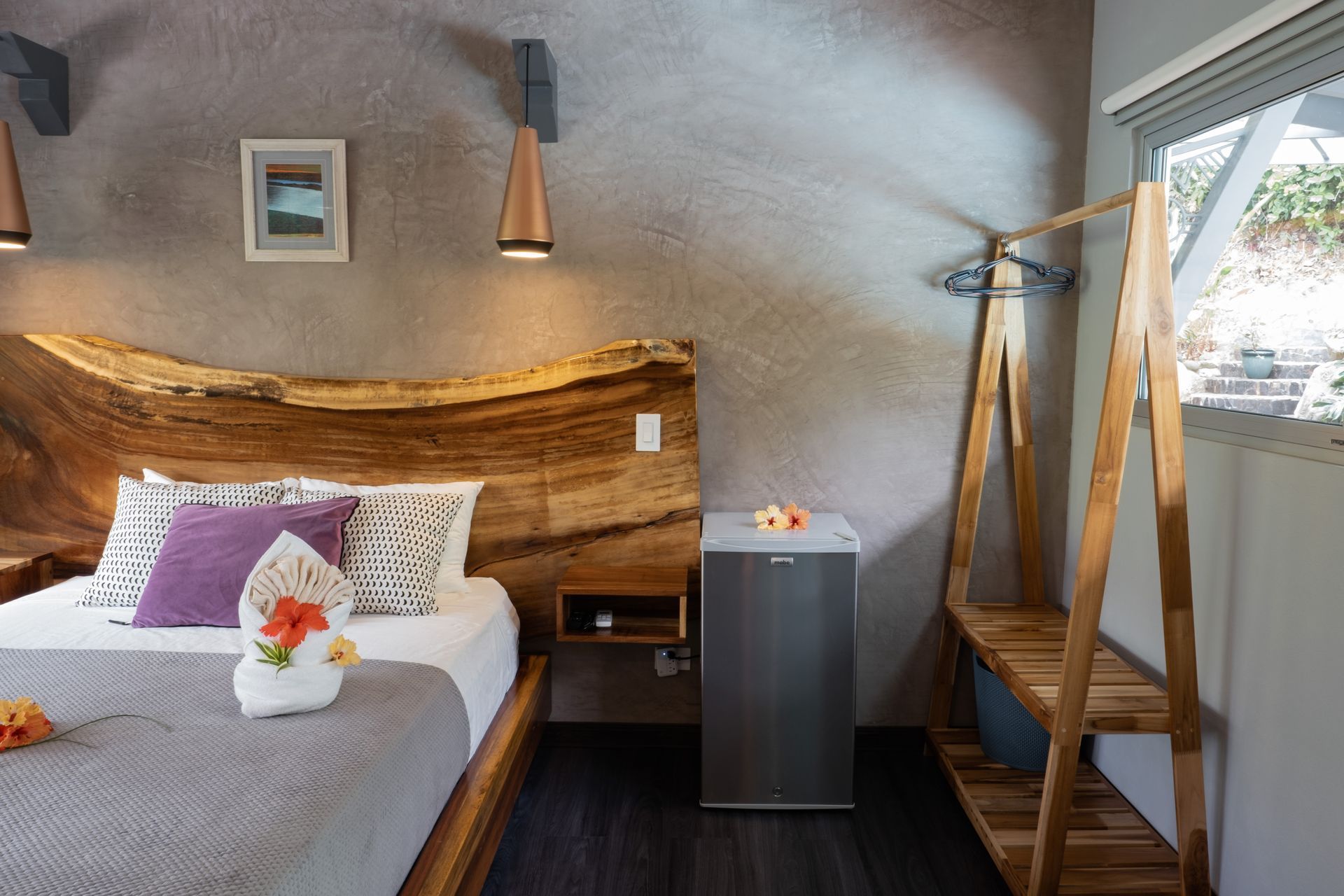 A bedroom with a live-edge wooden headboard, a white bed with a purple pillow, a small fridge, and a wooden clothes rack.