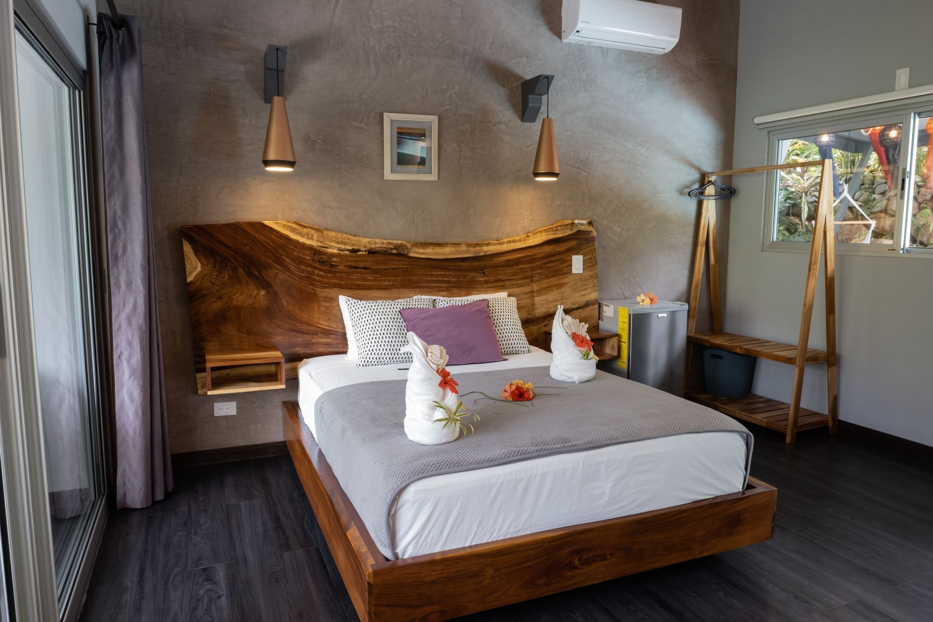A modern hotel room with a live-edge wooden bed frame, a grey bedspread, decorative towel swans, and a clothing rack.