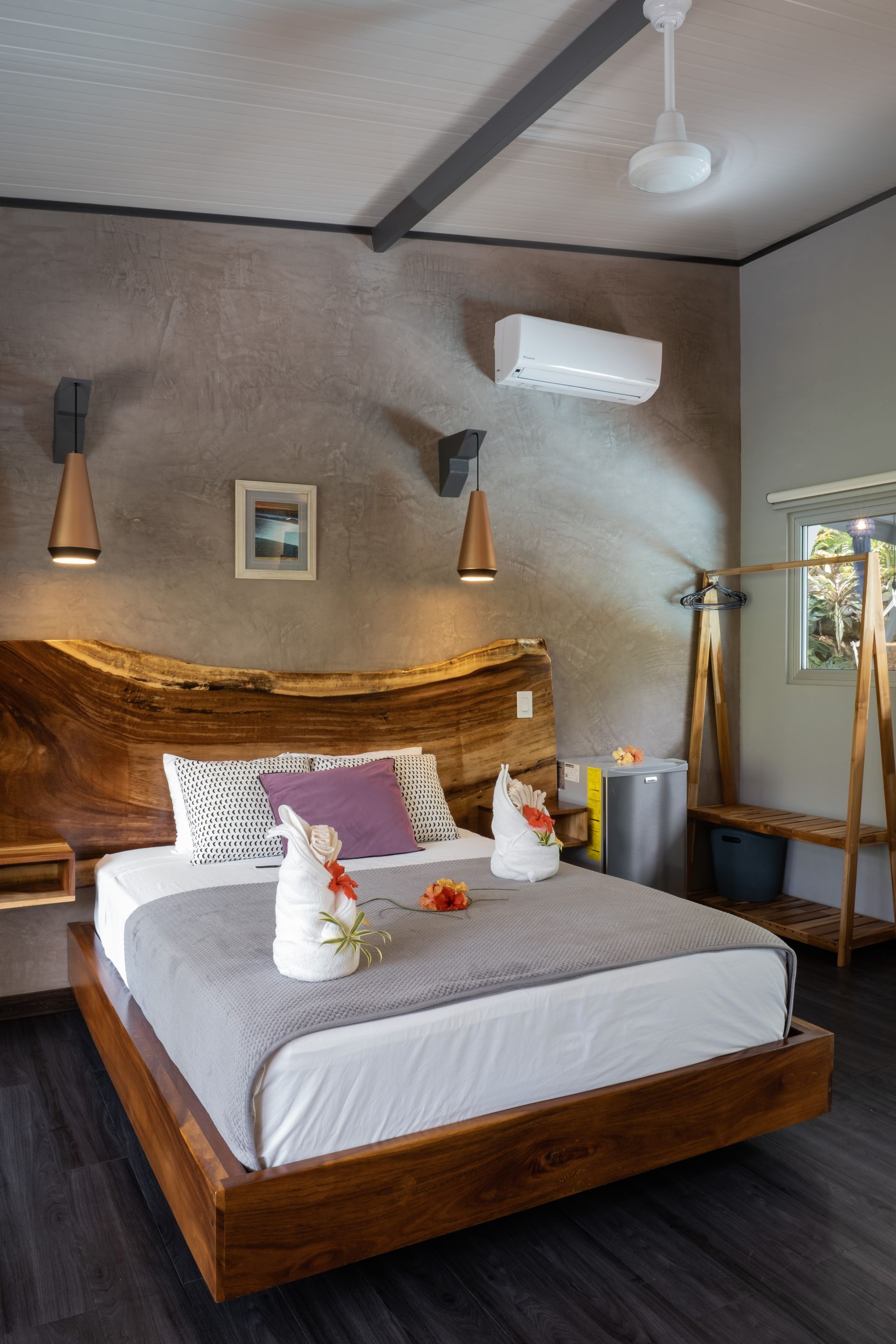 A modern bedroom features a live-edge wood headboard, a white bed with gray linens, two towel animals, and wall sconces.