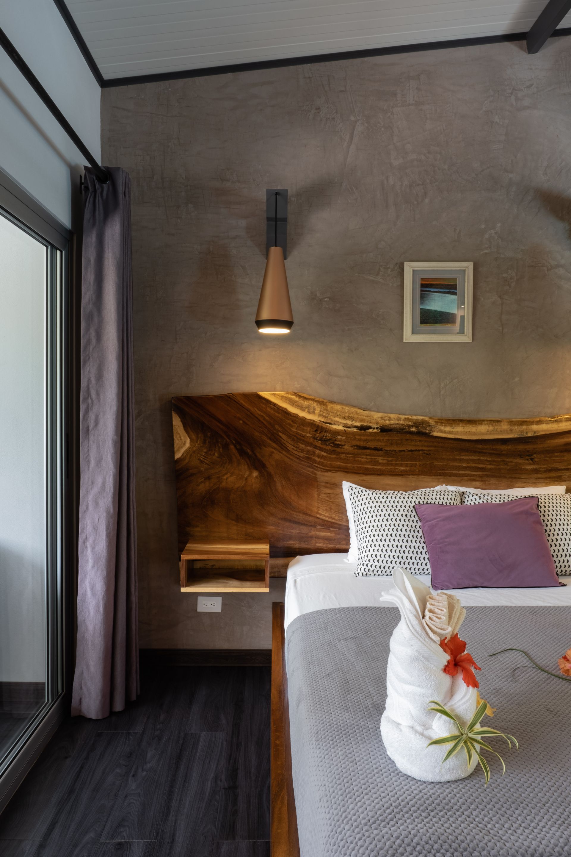 A hotel bedroom features a raw-edge wood headboard, a gray bed with a towel sculpture, and a contemporary wall lamp.