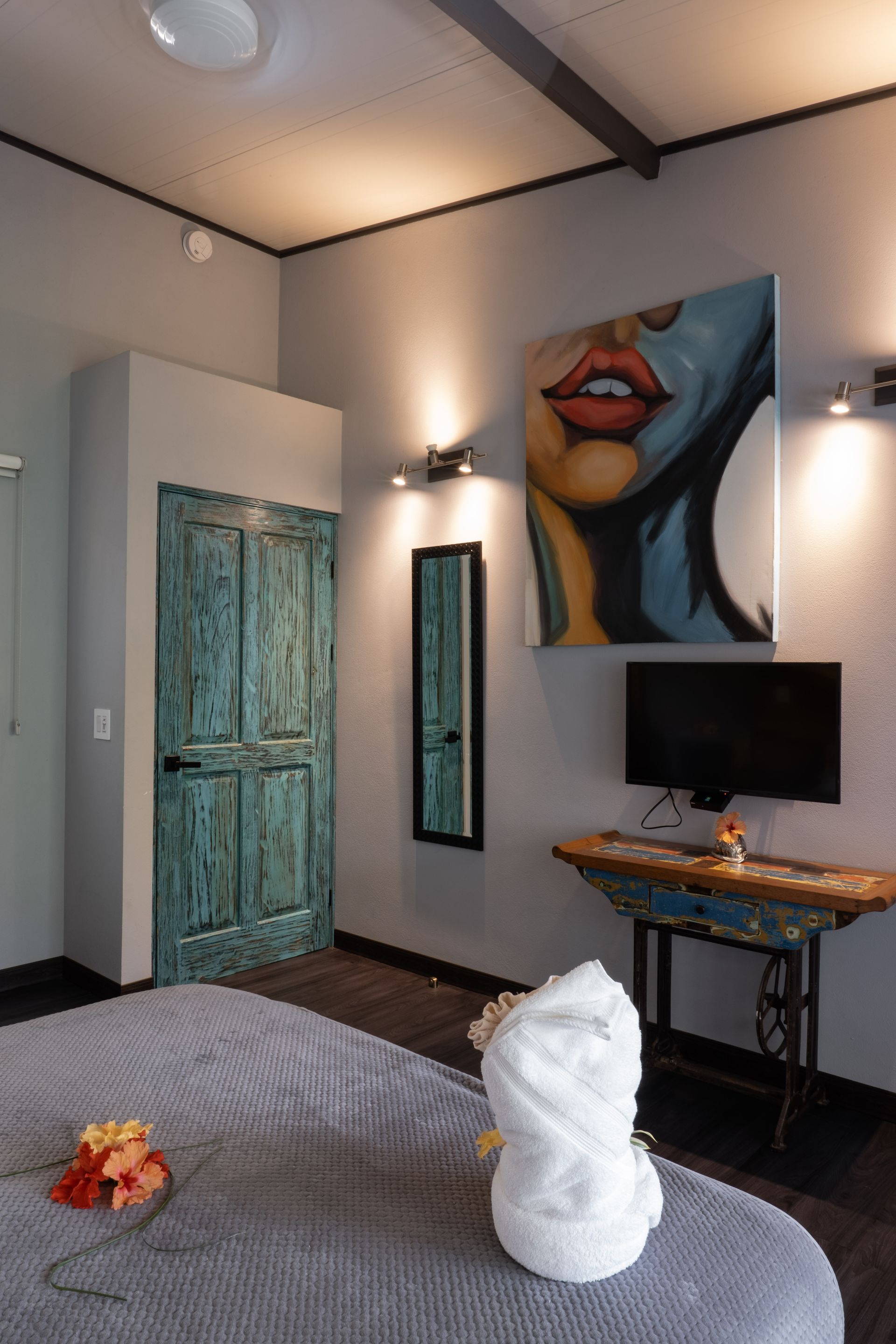 A hotel room with a textured teal door, a pop-art painting, a flat-screen TV on a vintage sewing machine table, and bed.
