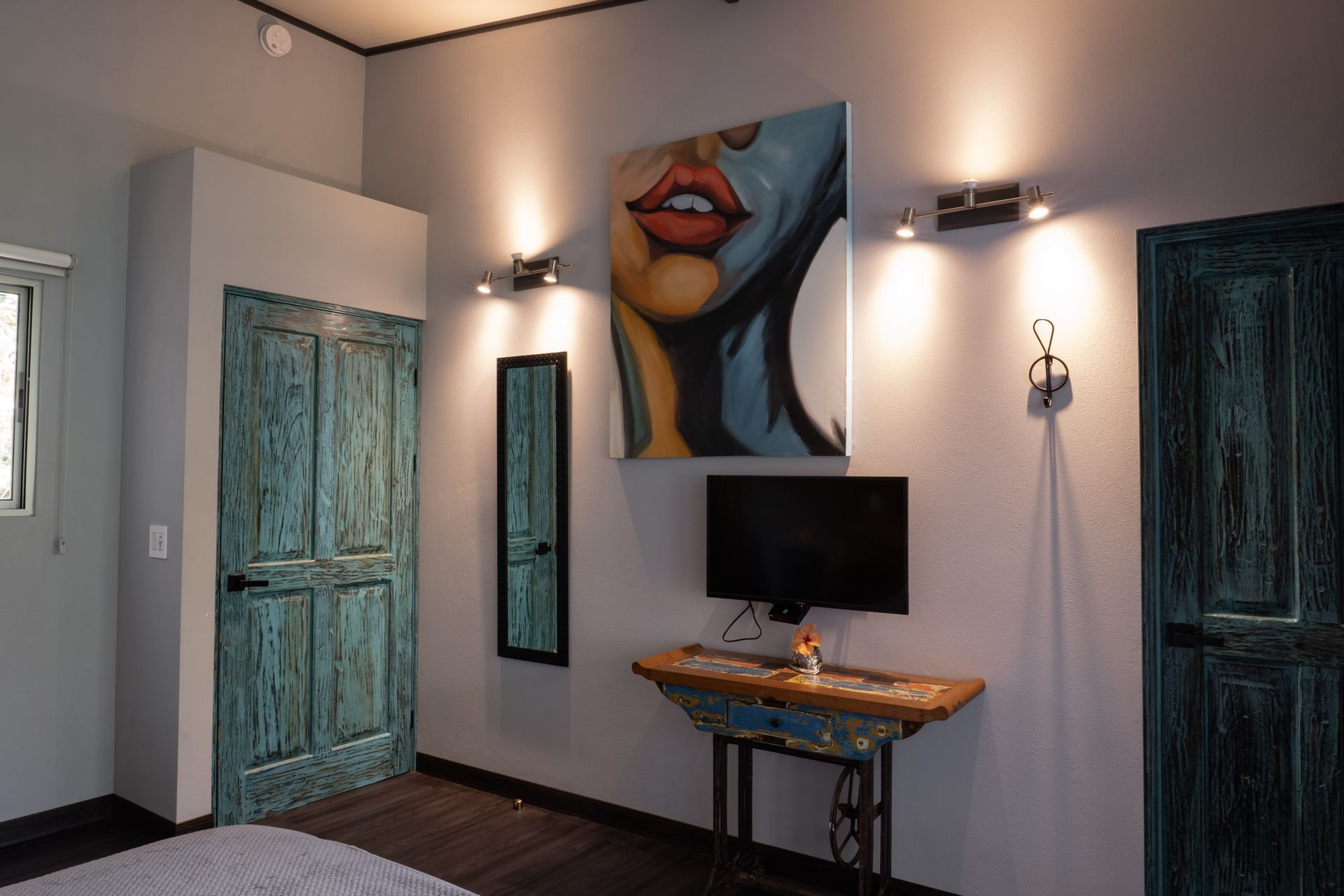A bedroom wall with two distressed teal doors, a painting of a face, a small television, and a vintage sewing table.