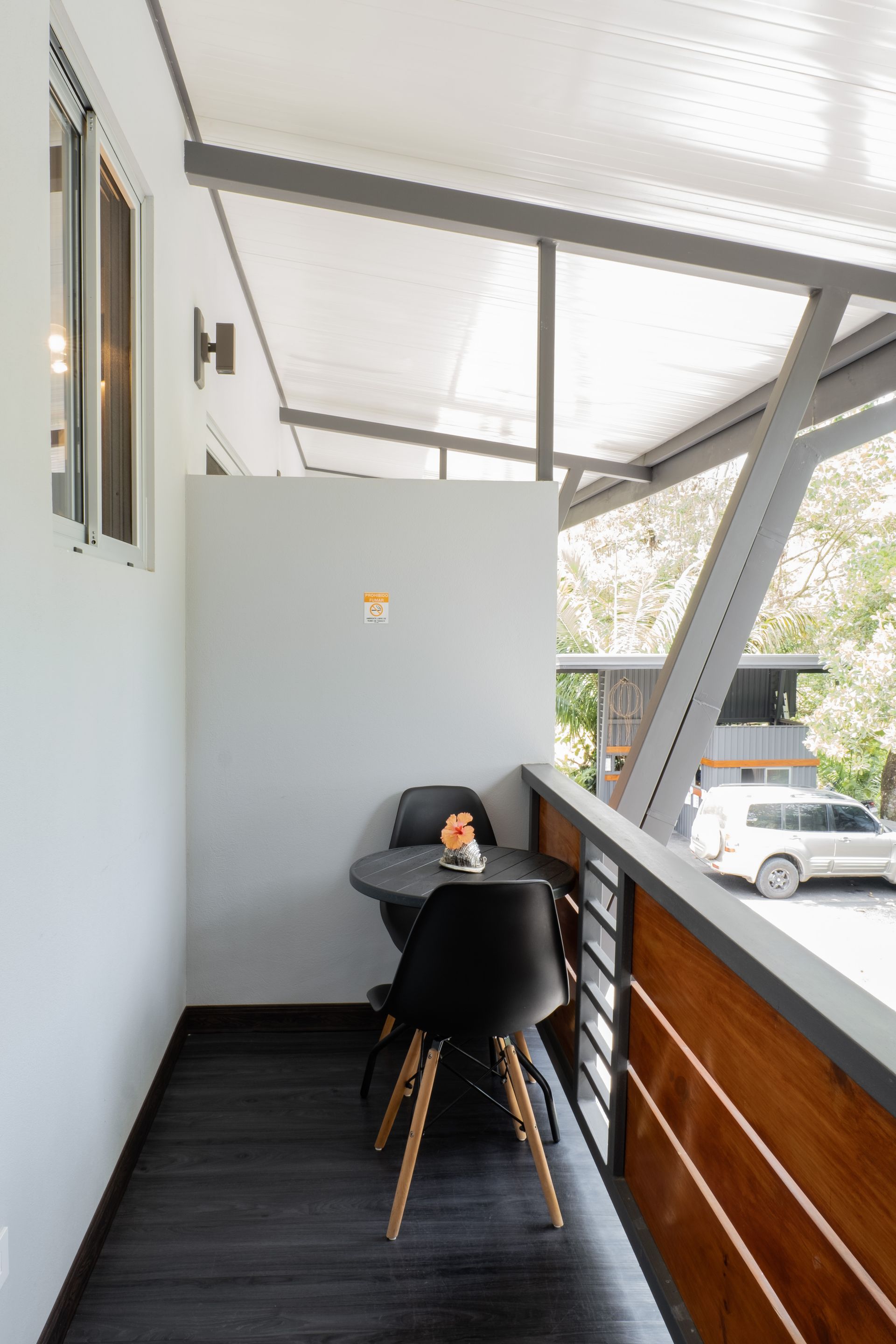A covered balcony featuring a small round table with two black modern chairs, dark flooring, and a wooden side railing.