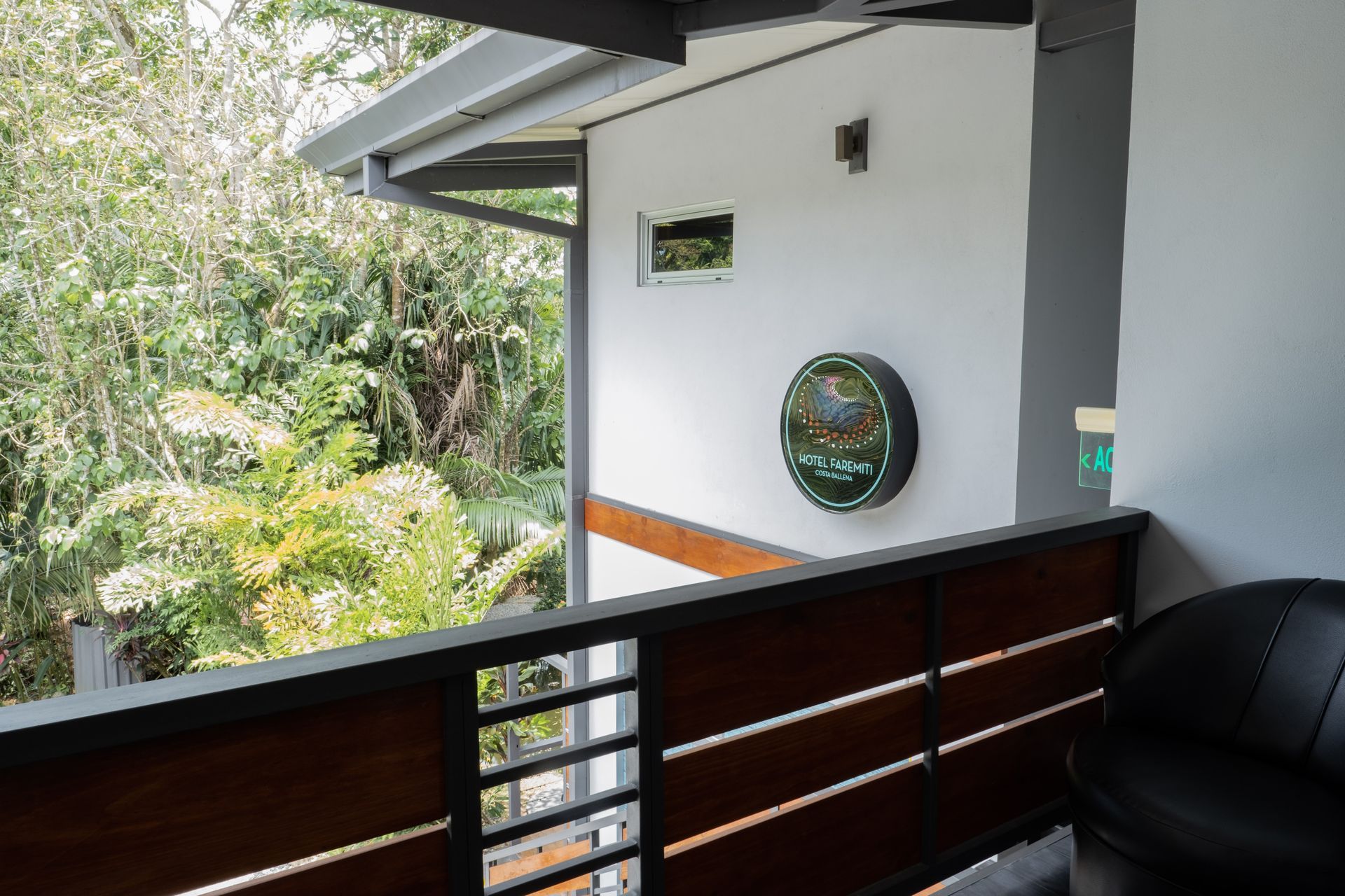 A balcony with a wooden railing overlooks lush trees, with a circular Heineken sign mounted on a white exterior wall.