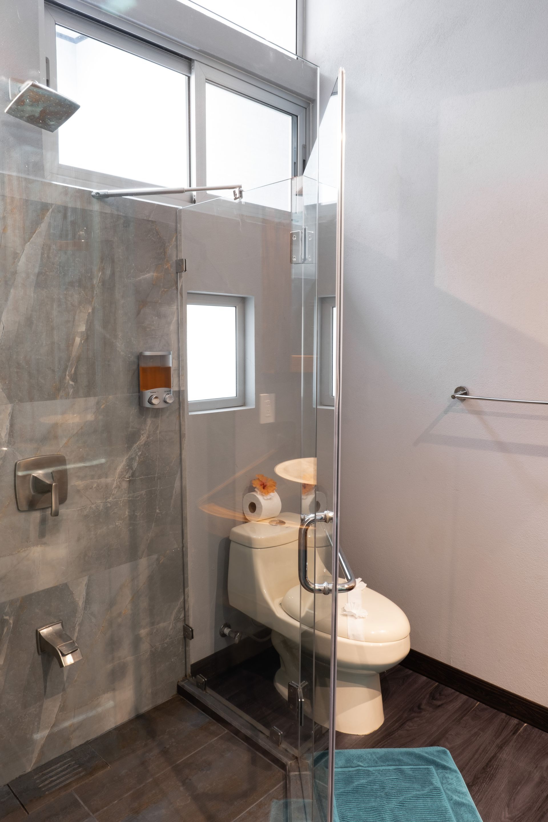A modern bathroom featuring a glass-enclosed shower with gray tiled walls next to a white toilet on dark wood-look flooring.