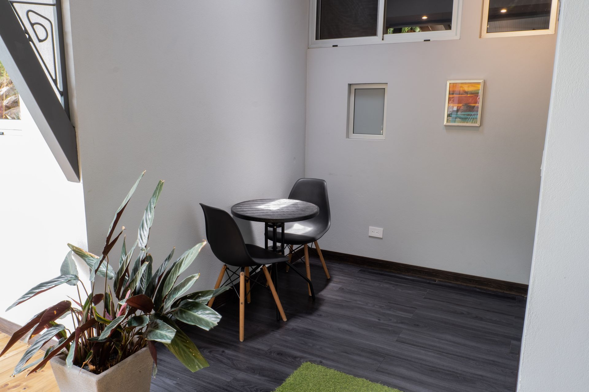 A small, modern seating area with a round table and two black chairs, next to a large potted plant on a dark wood floor.