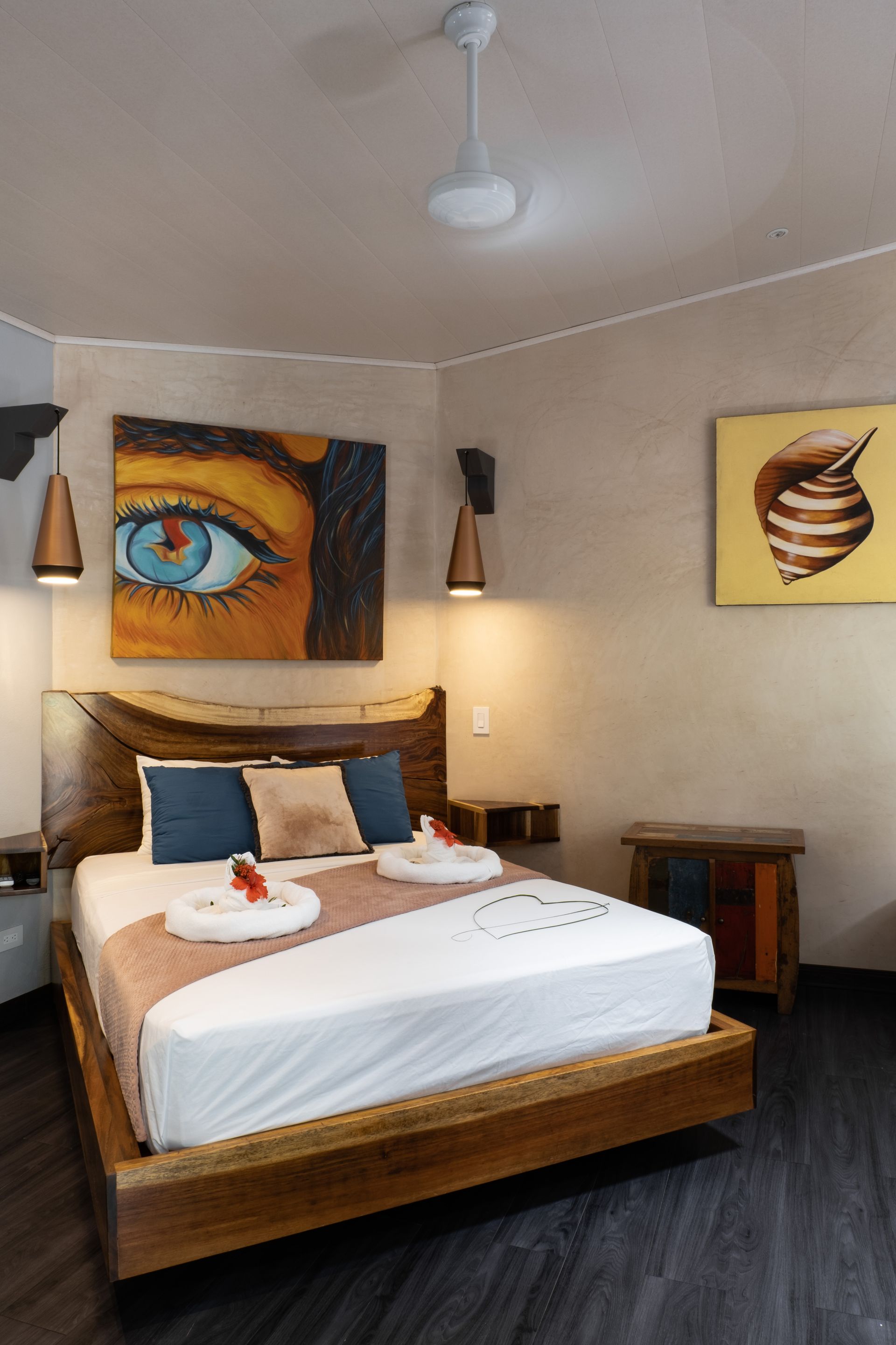 A modern bedroom featuring a bed with white linens, a wooden frame, and a large eye painting above the headboard.