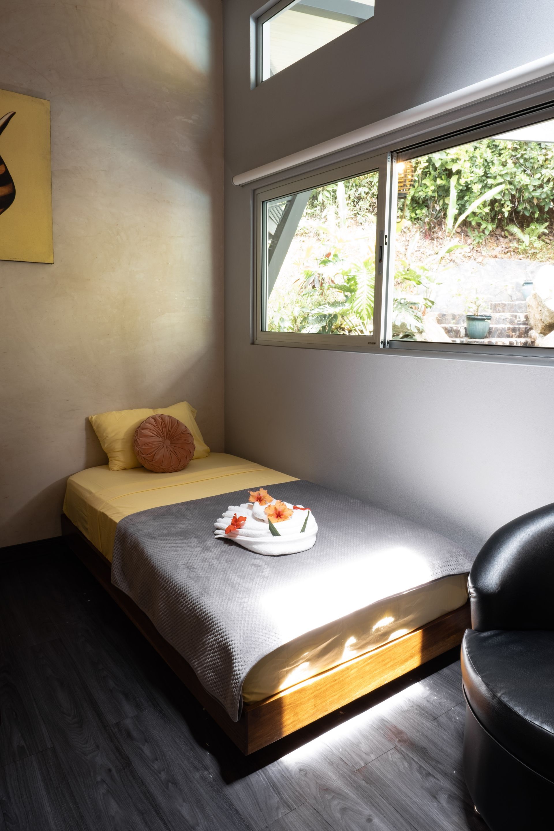 A twin bed with yellow bedding, a gray blanket, and floral accents sits in a sunlit room next to a window and chair.