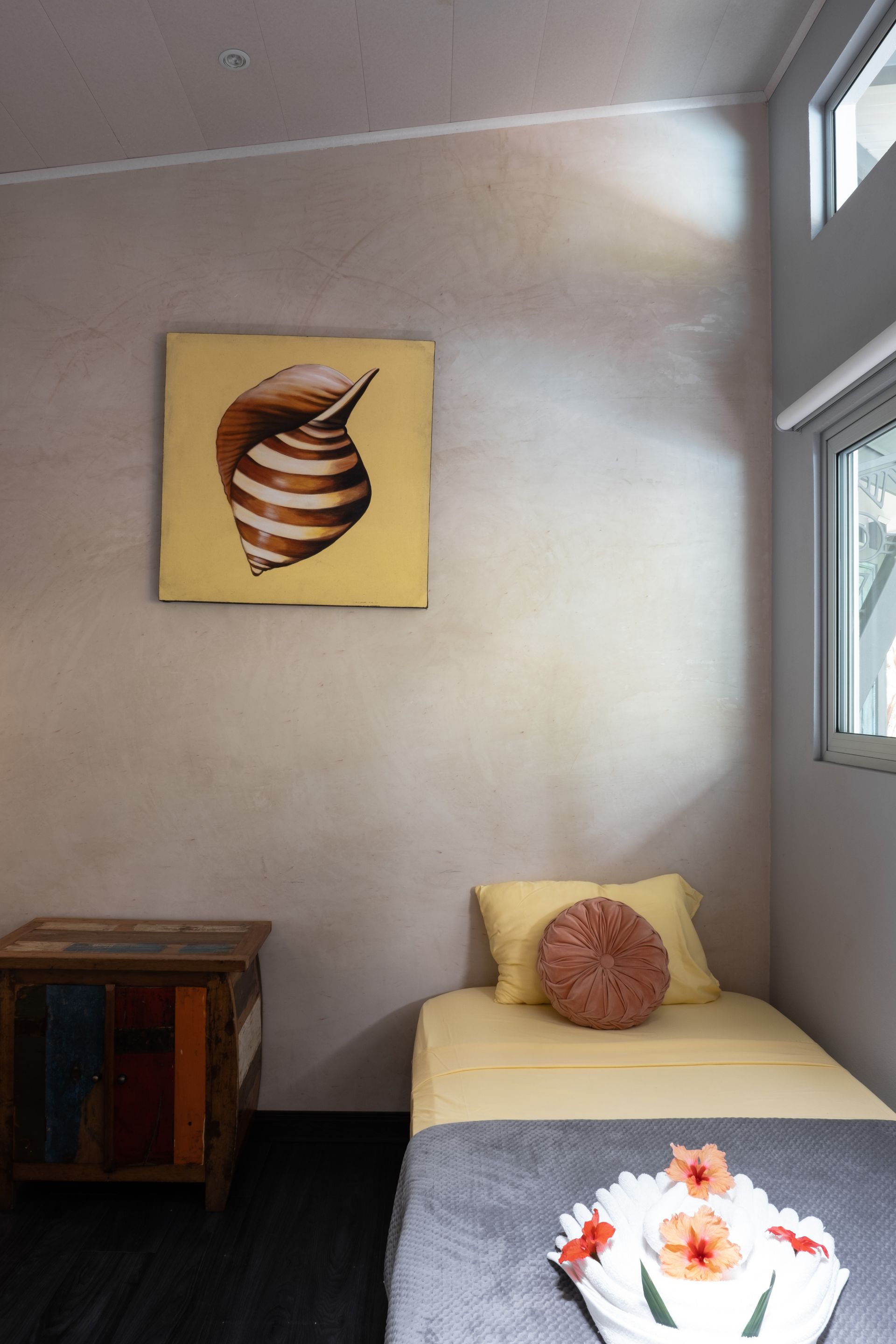 A small bedroom with a single bed, a wooden nightstand, and a framed seashell painting on a neutral wall.